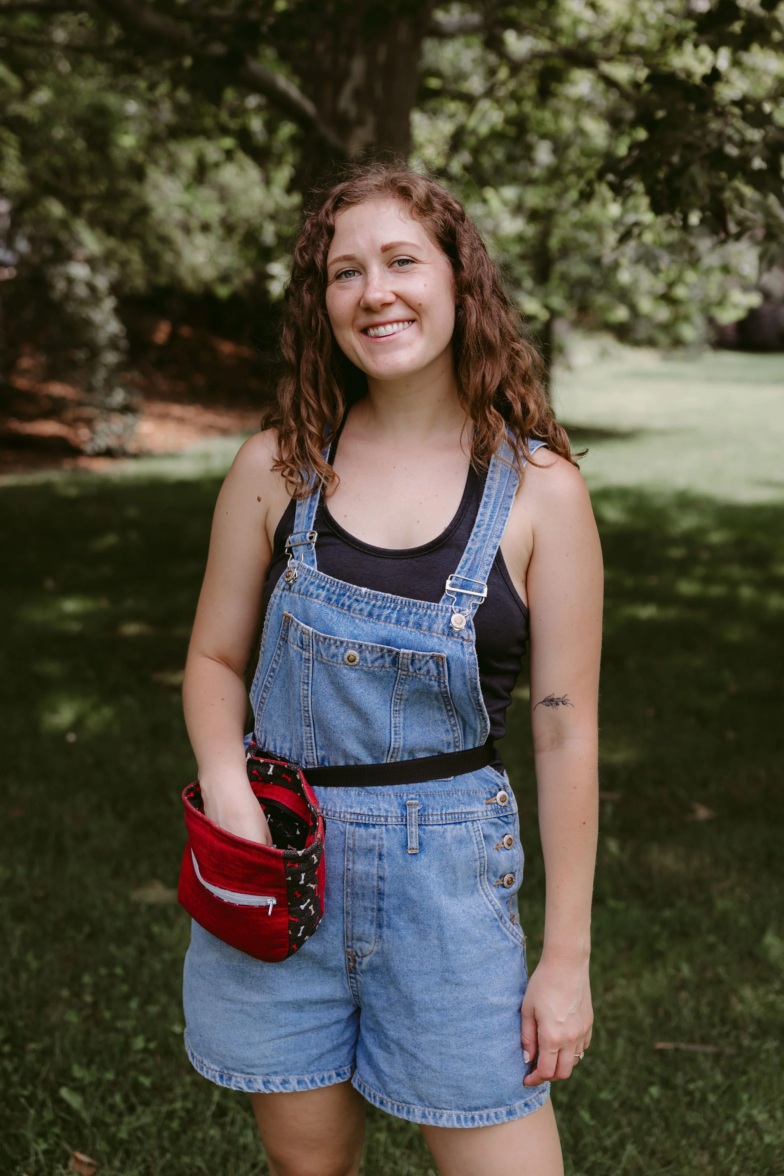 A young woman with curly brown hair smiling outdoors, standing on grass with trees in the background, wearing a black tank top, denim overalls, and holding a red and black bag.