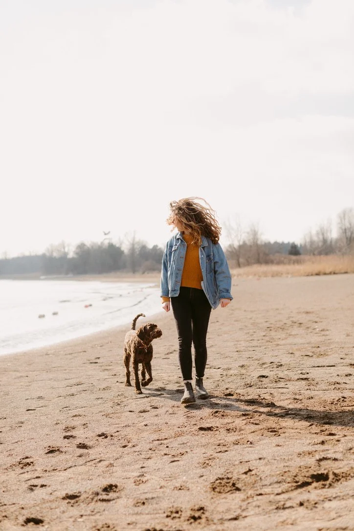 A woman walking a dog on a beach during daytime, with trees and water in the background.