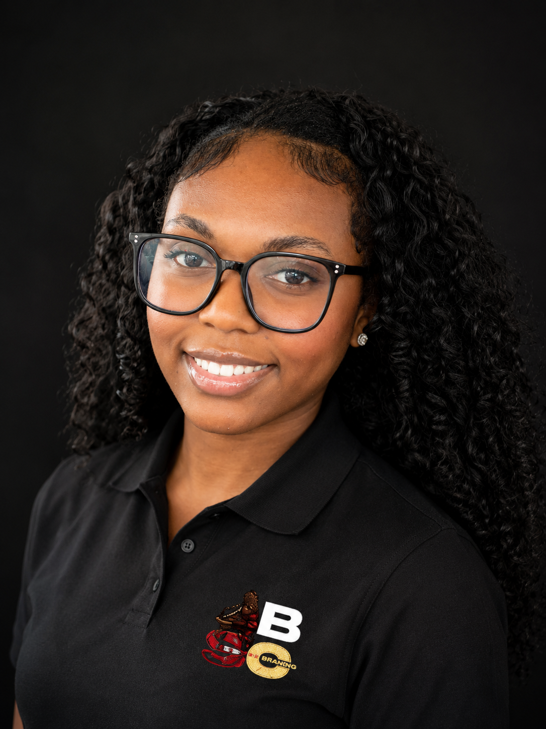 A young woman with curly black hair and glasses, wearing a black collared shirt with various embroidered logos, smiling against a black background.
