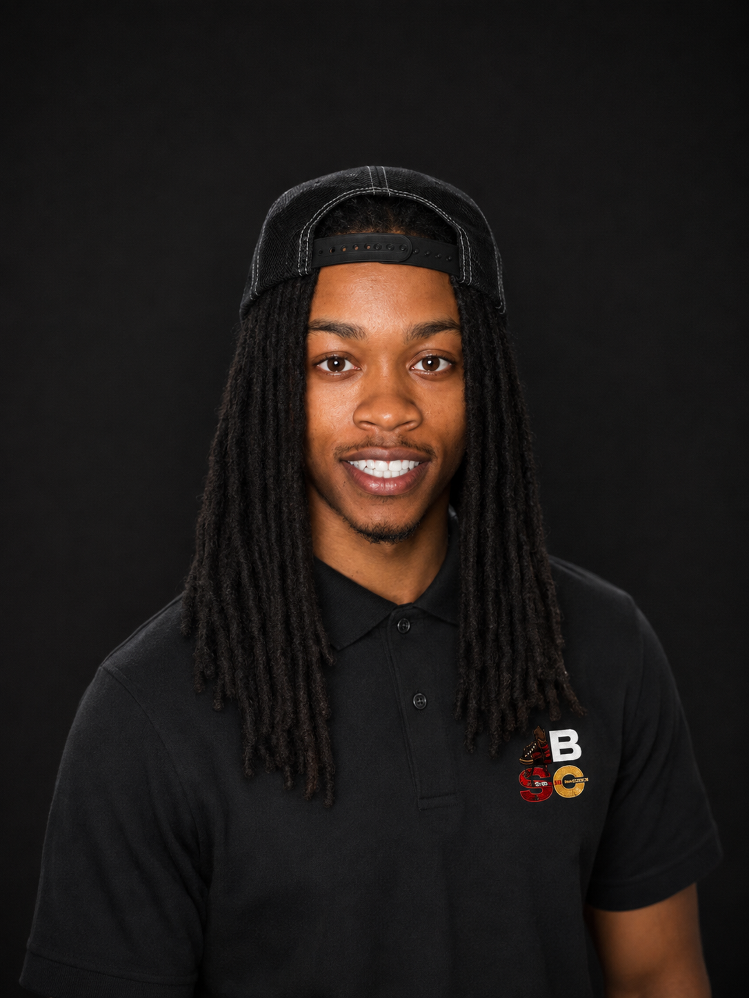 Portrait of a smiling young man with dreadlocks wearing a black baseball cap backwards and a black polo shirt with a logo on the chest, set against a black background.