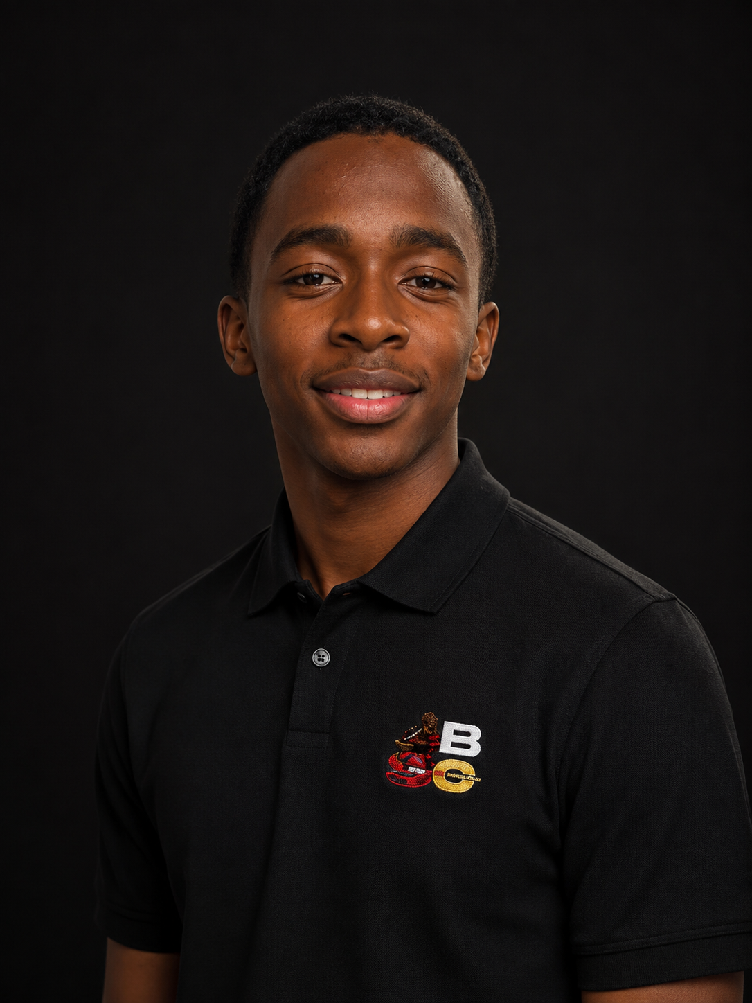 Young man with short curly hair smiling at camera, wearing a black polo shirt with colored logo patches on the chest, against a black background.