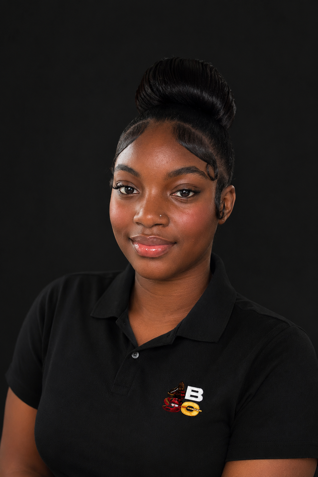 Portrait of a young African American woman with a high, styled bun, wearing a black polo shirt with a logo that includes a cupcake and the letters 'B' and 'G', against a plain black background.