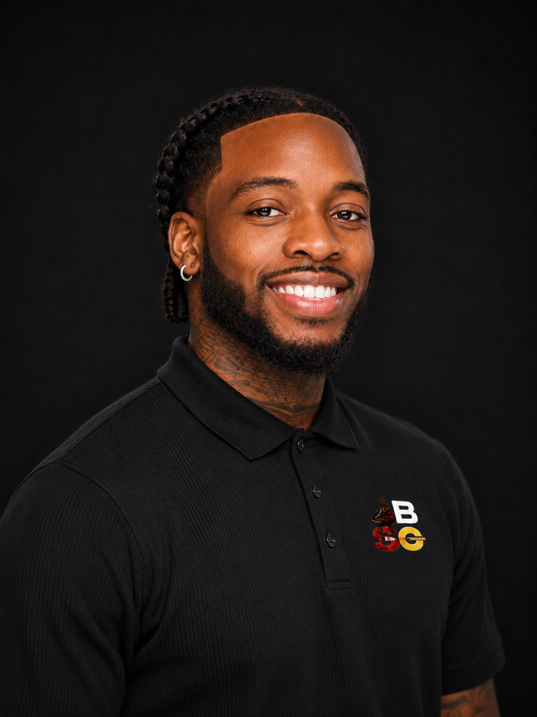 Young man with braided hair, beard, and earrings smiling in front of a black background, wearing a black polo shirt with a logo featuring a butterfly, a bee, and the letters B, S, and G.