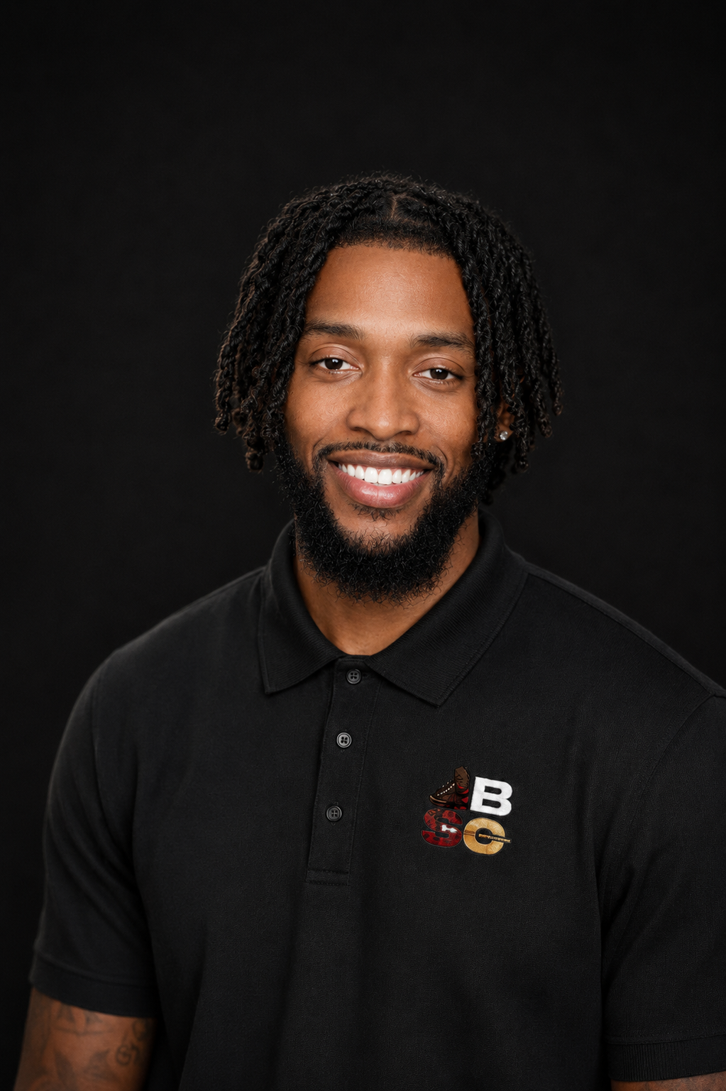 A smiling man with dark curly hair and a beard, wearing a black polo shirt with a logo on the chest, against a black background.