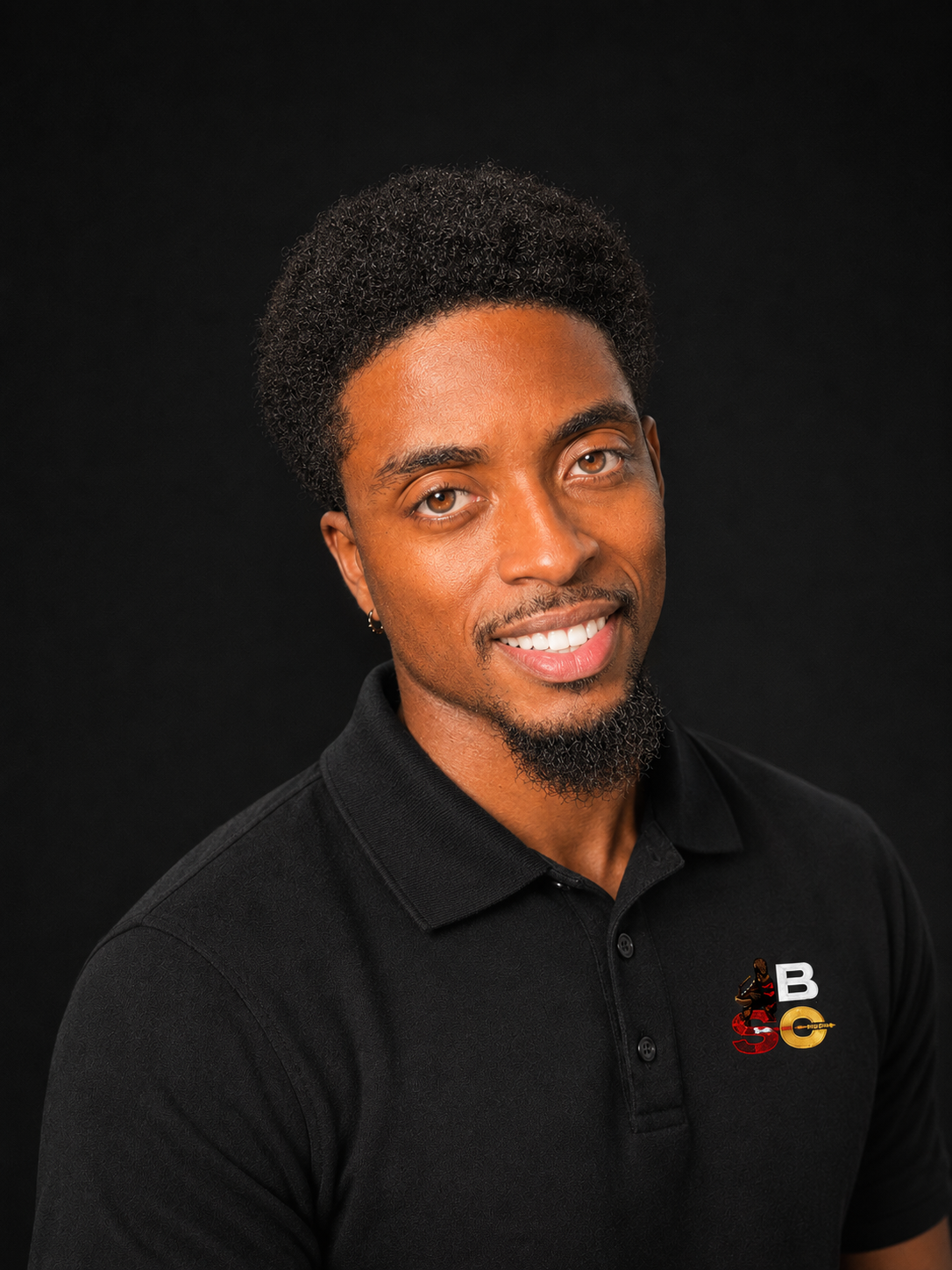 Portrait of a smiling young man with curly hair and a goatee, wearing a black polo shirt with a logo featuring a woman, the letters B and G, and a gold circular element, against a black background.