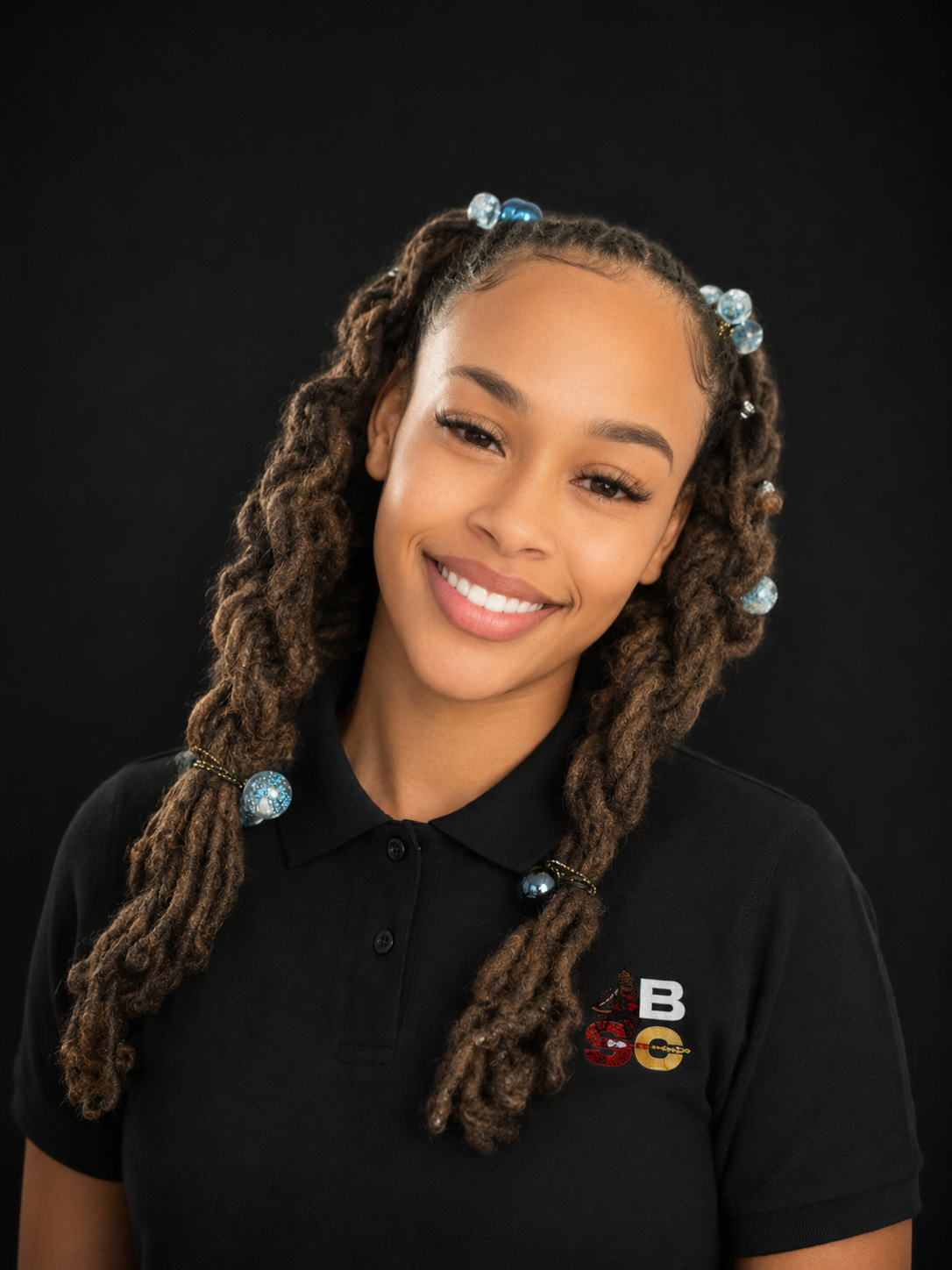 A young woman with styled dreadlocks decorated with blue and silver beads, smiling and wearing a black polo shirt with a logo on it, against a black background.
