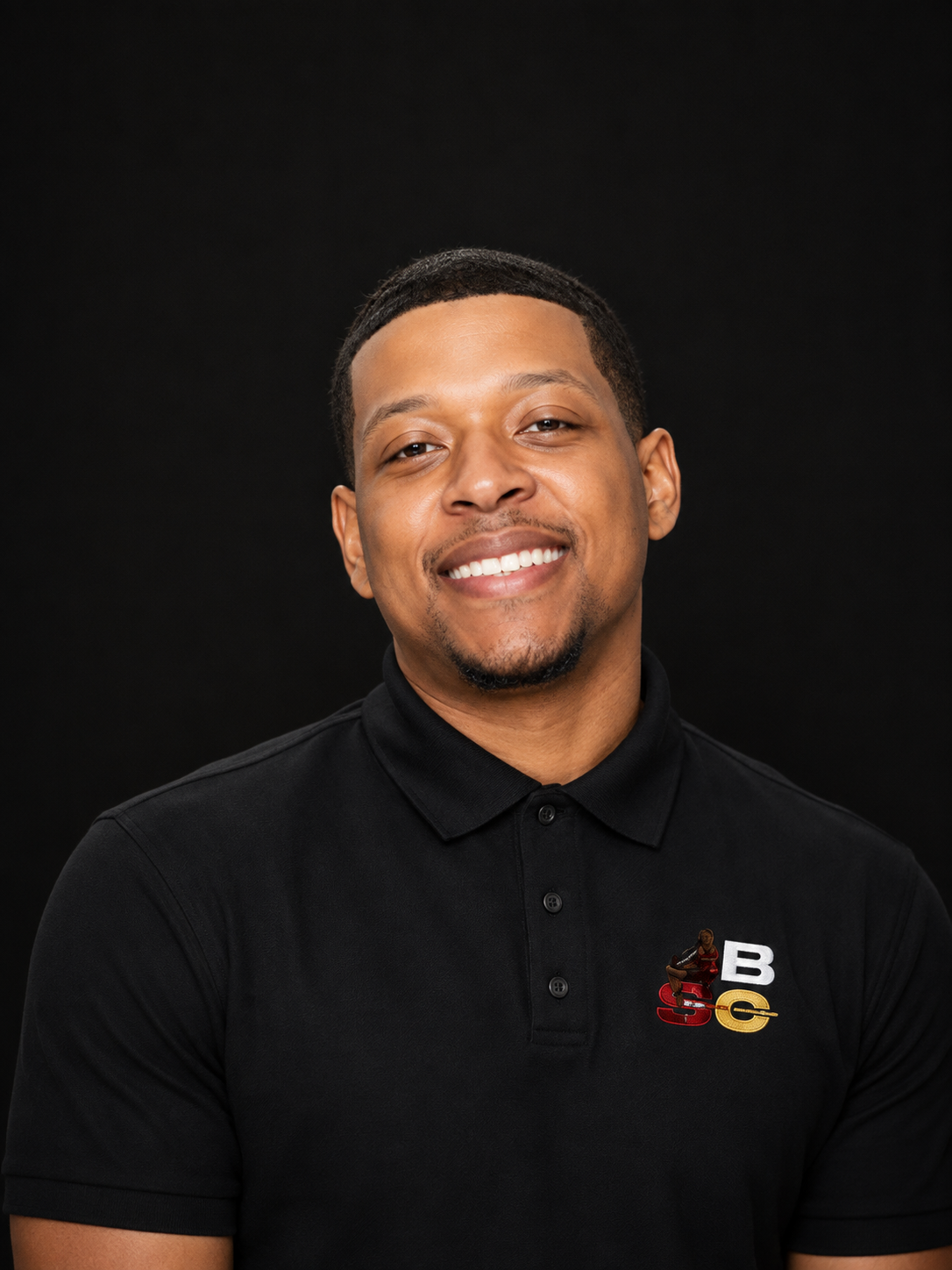 A smiling man with short black hair and a goatee wearing a black polo shirt with a logo on the chest, standing against a black background.