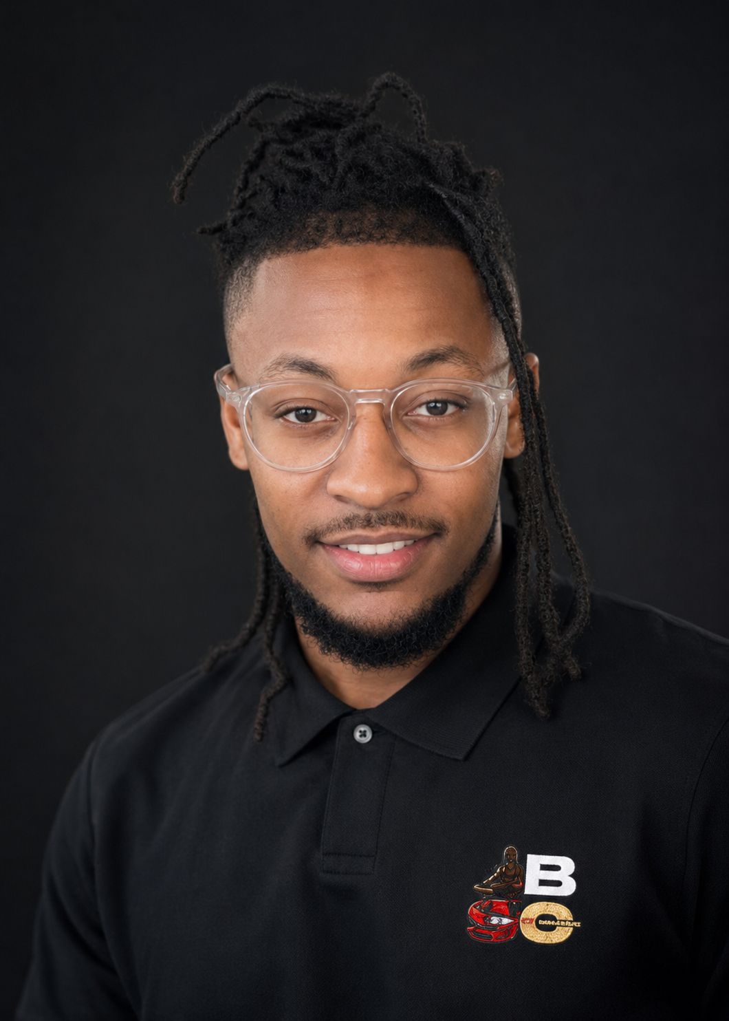 A young man with dreadlocks wearing clear glasses and a black polo shirt with a logo featuring a figure, car, and letters B and C, posing against a dark background.