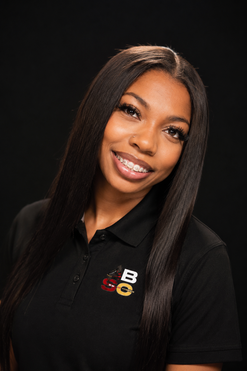 A young woman with long straight black hair, smiling, wearing braces and a black polo shirt with a logo.