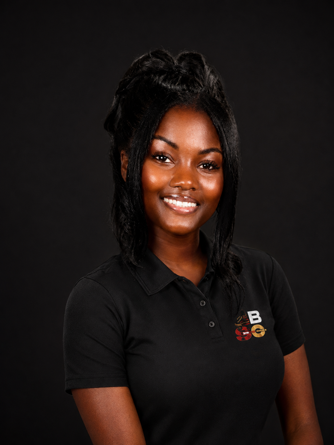 Woman with dark skin and styled black hair smiling, wearing a black collared shirt with a logo on the chest, against a dark background.
