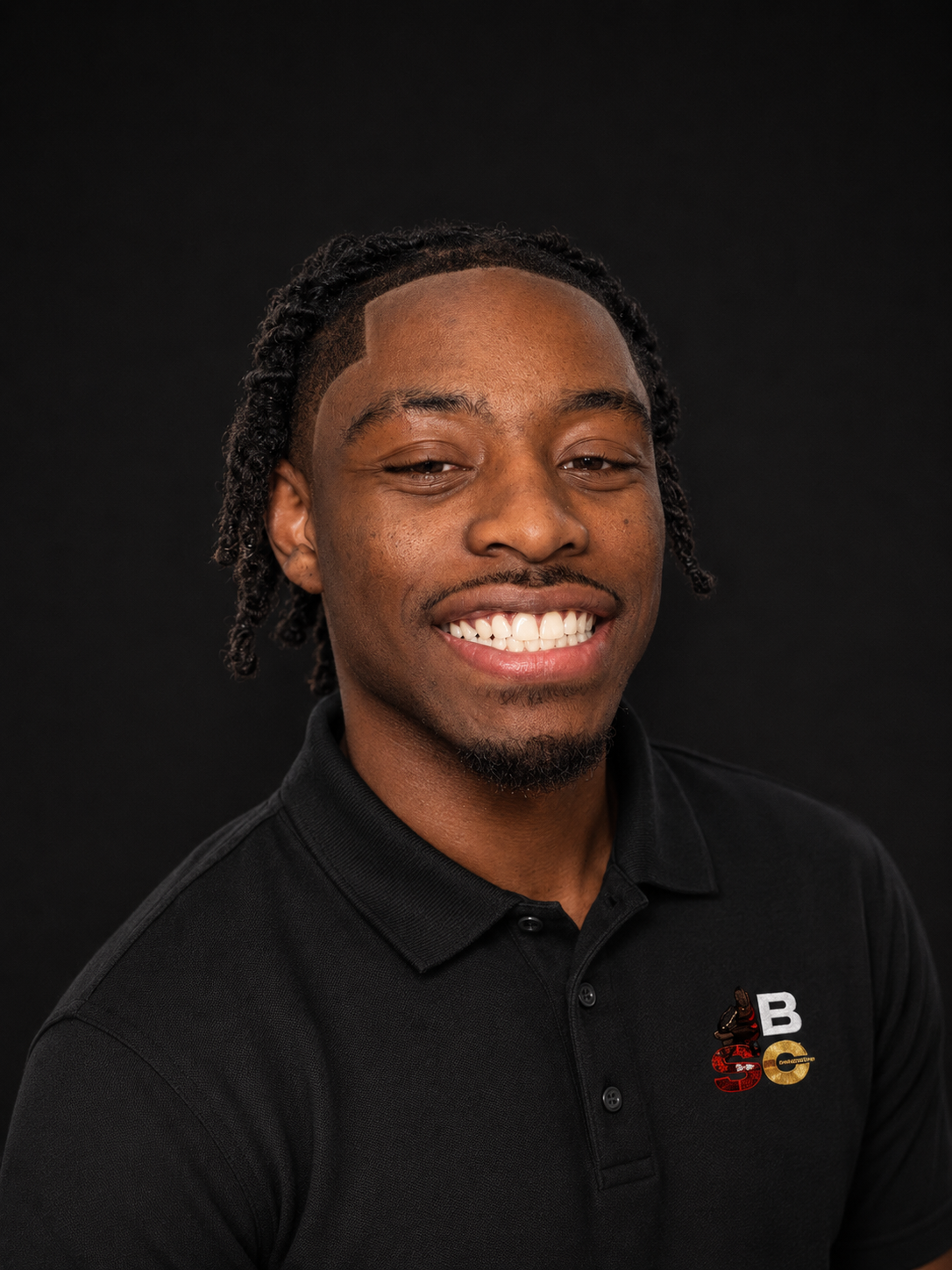 Headshot of a smiling young man with short curly hair and a goatee, wearing a black polo shirt with a logo on the chest, against a dark background.