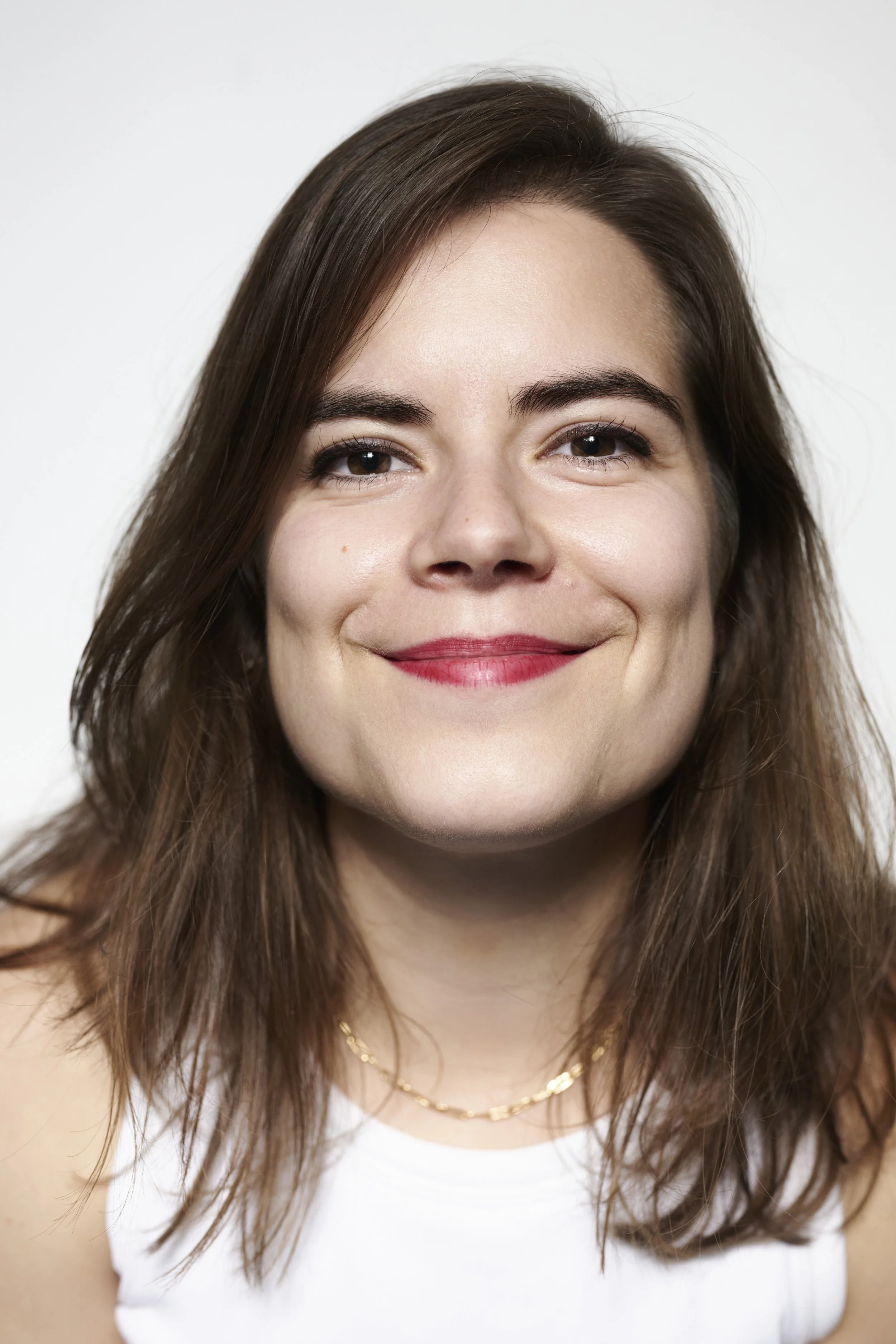Close-up of a young woman with medium-length brown hair, wearing a white top, gold necklace, and smiling against a plain background.