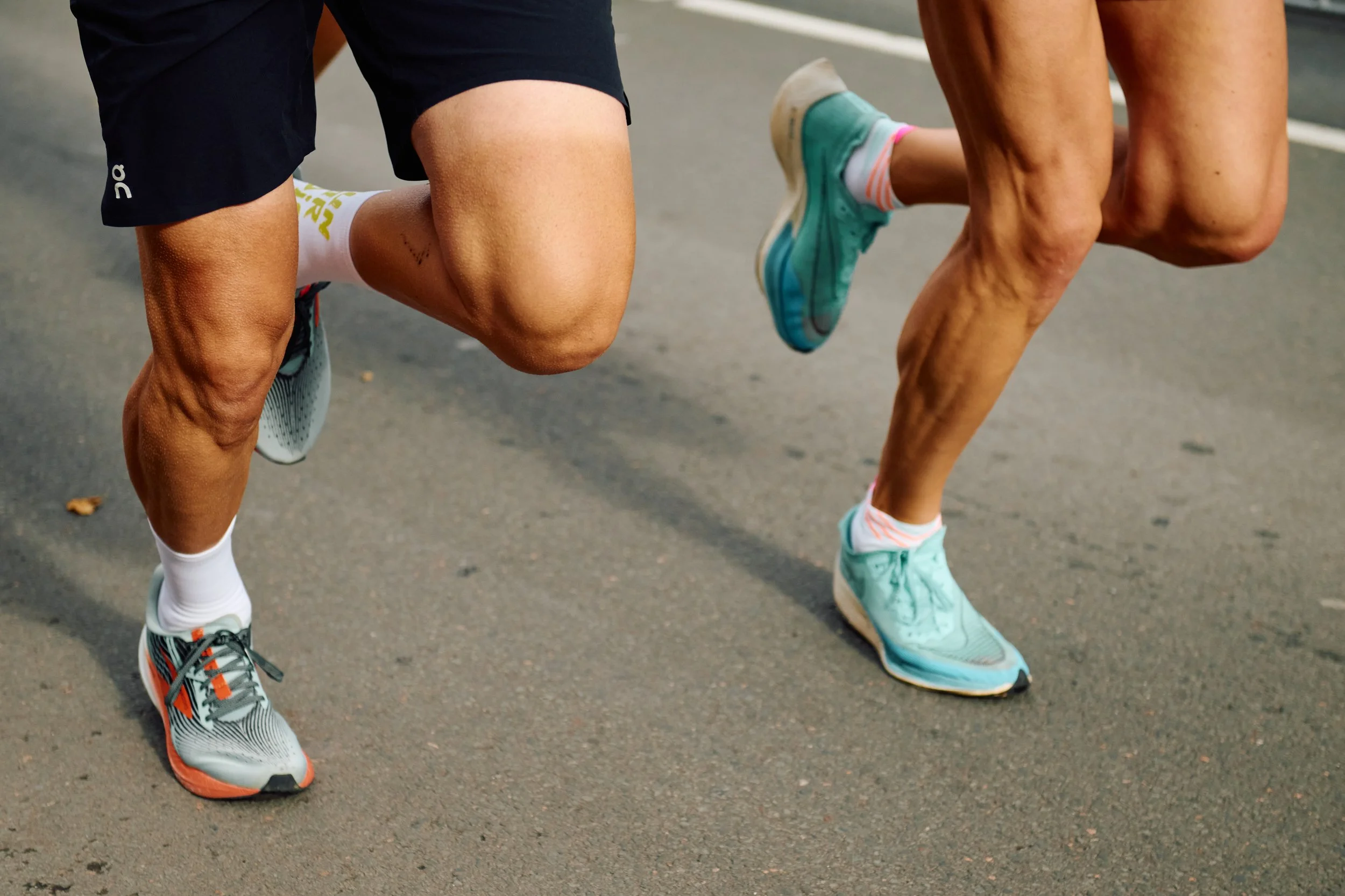 Close-up of two runners' legs and feet as they race on a paved road, wearing athletic shoes and shorts.