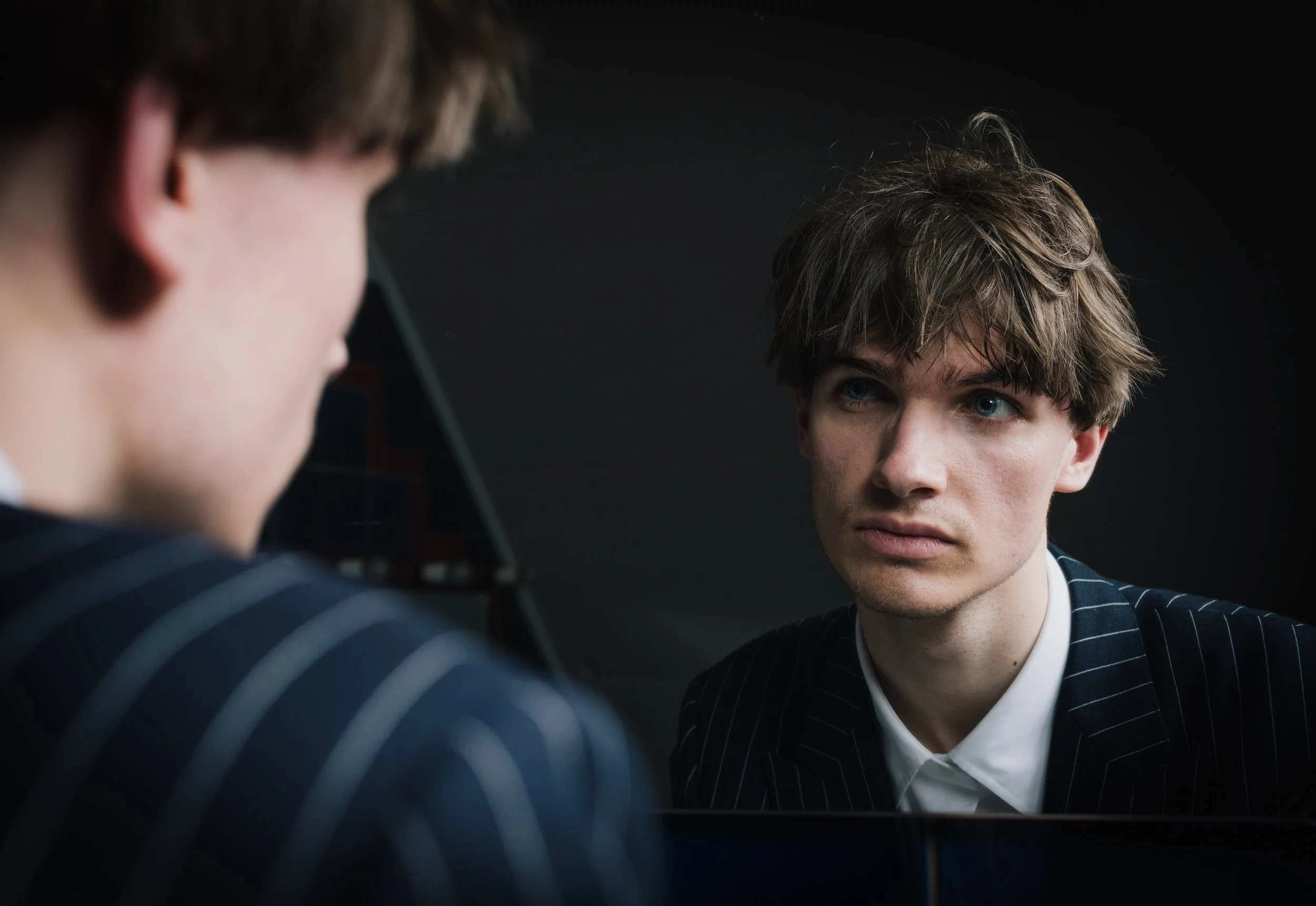 A young man with brown, messy hair looking at his reflection in a mirror, wearing a dark pinstripe suit and a white shirt, set against a dark background.