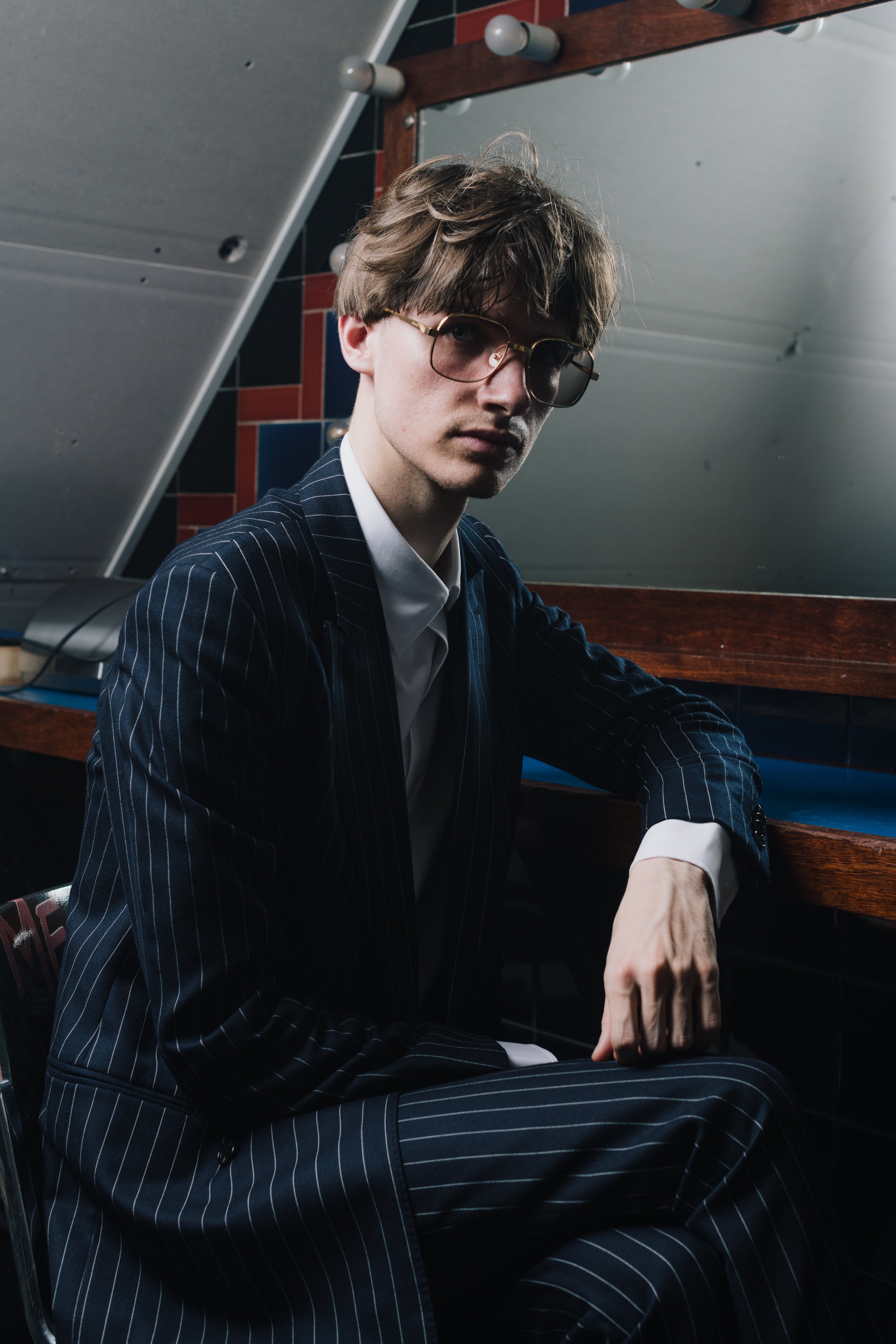 Young man with glasses in a pinstripe suit sitting in a dimly lit room with a wooden counter and mirror behind him.