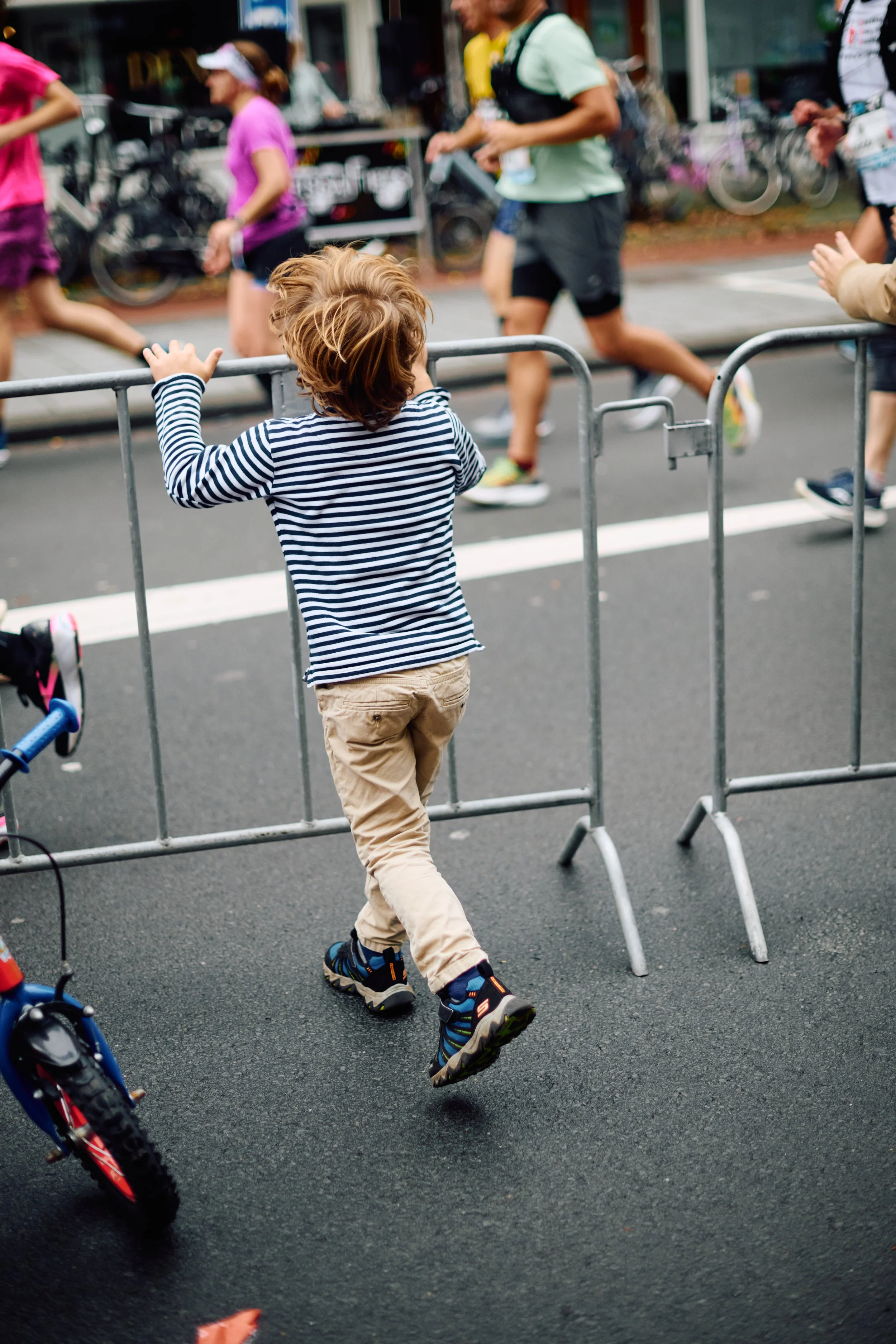 A young boy with red hair wearing a striped shirt and beige pants, holding onto a metal barricade at a street event with runners passing by.