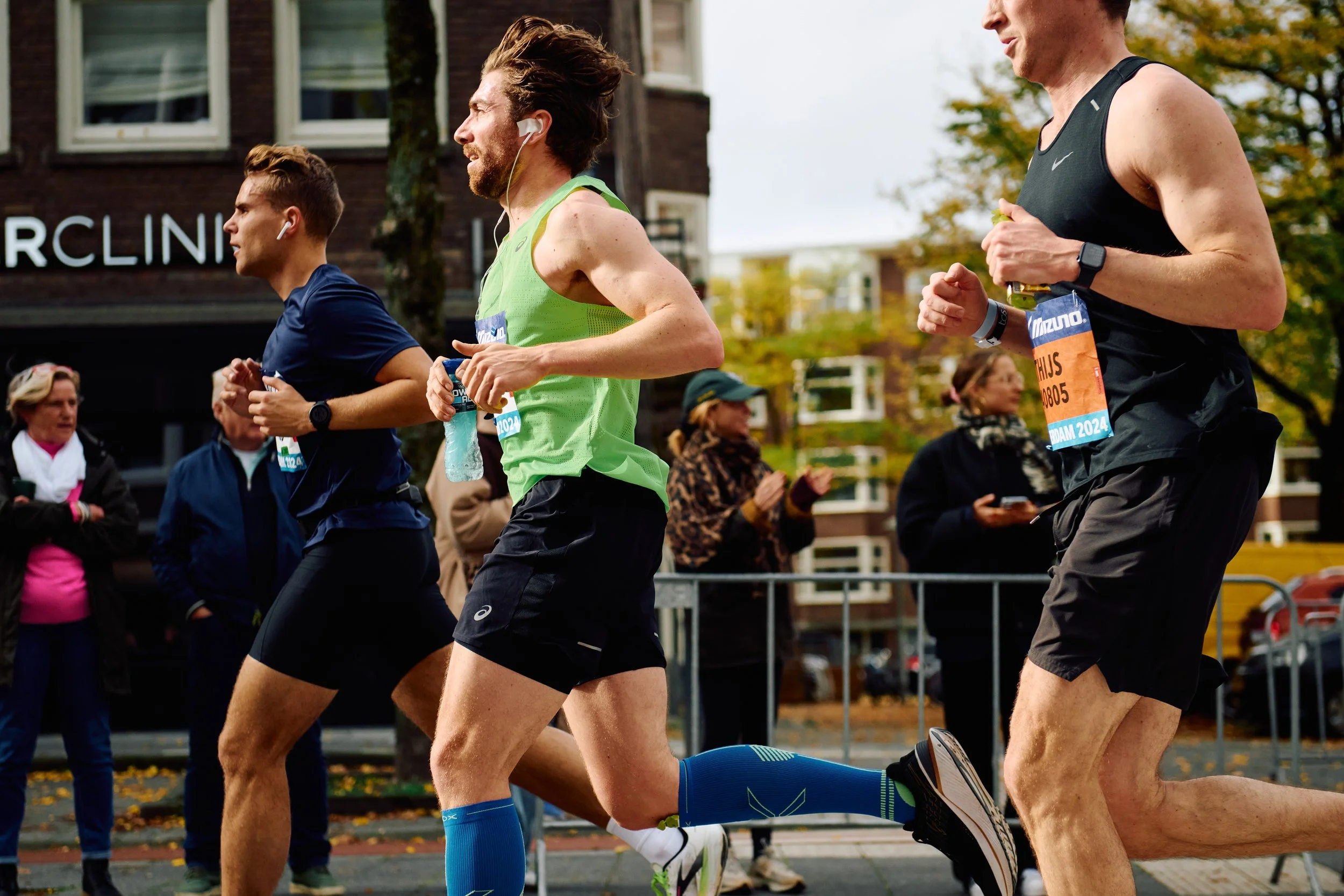 Three male runners participating in a marathon race, running outdoors on a city street with spectators watching in the background.