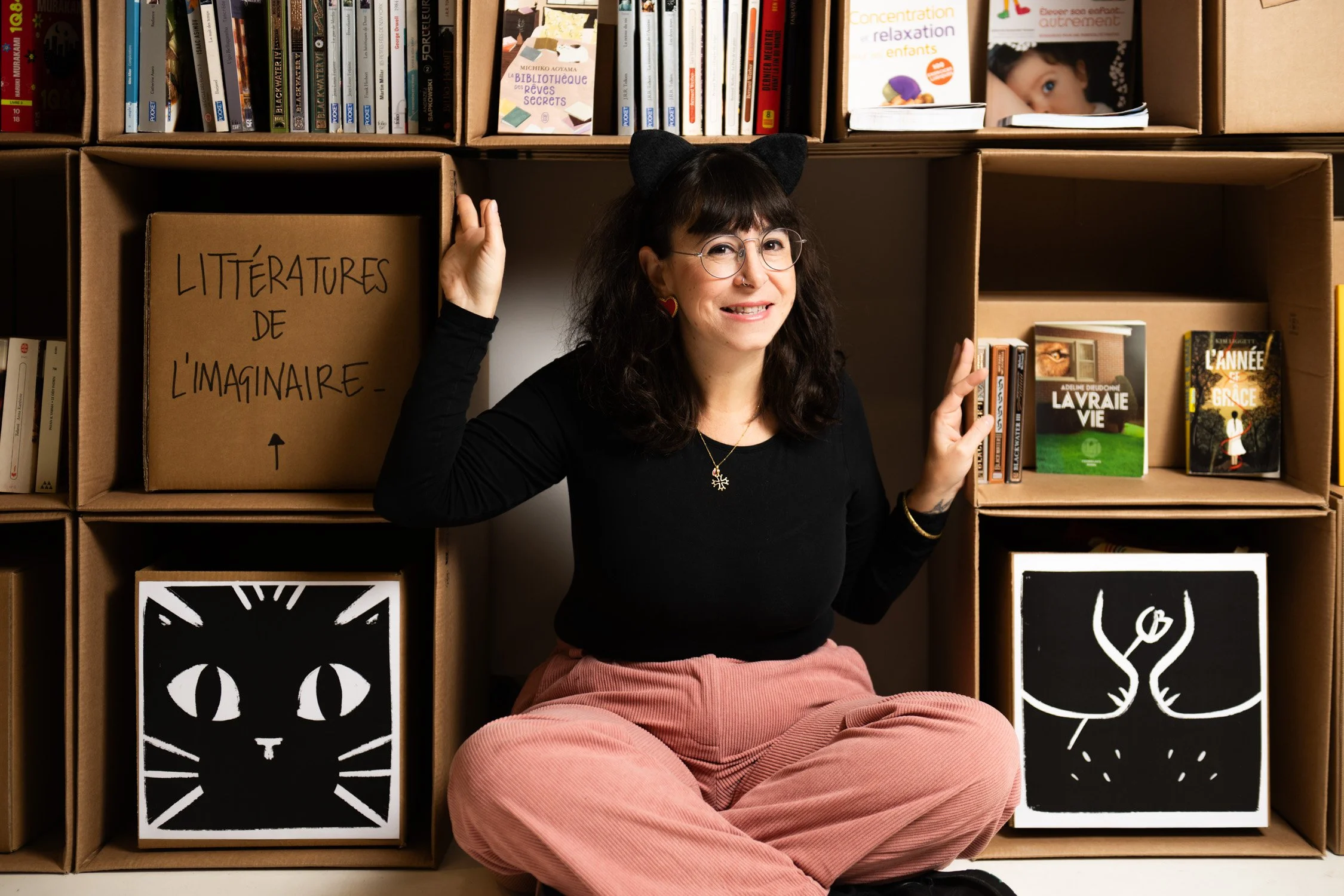 A woman with glasses, cat ears headband, and black top, sitting on the floor in front of wooden shelves filled with books and cardboard boxes. She is smiling and holding a box labeled "Littératures de l'imaginaire" with a black and white cat illustra