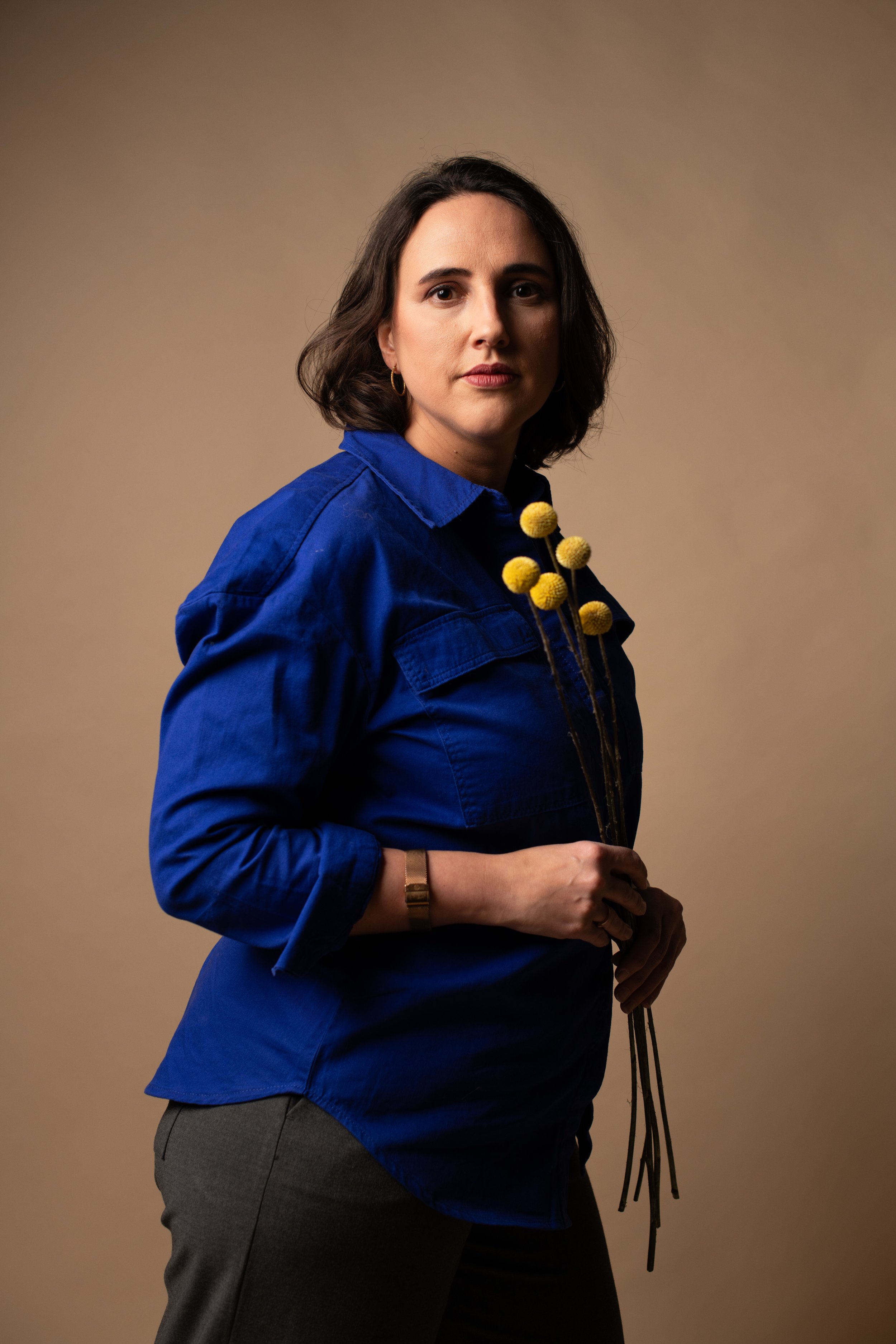 A woman with shoulder-length brown hair wearing a royal blue shirt and gray pants, holding a small bouquet of yellow billy buttons flowers, standing against a plain beige background.
