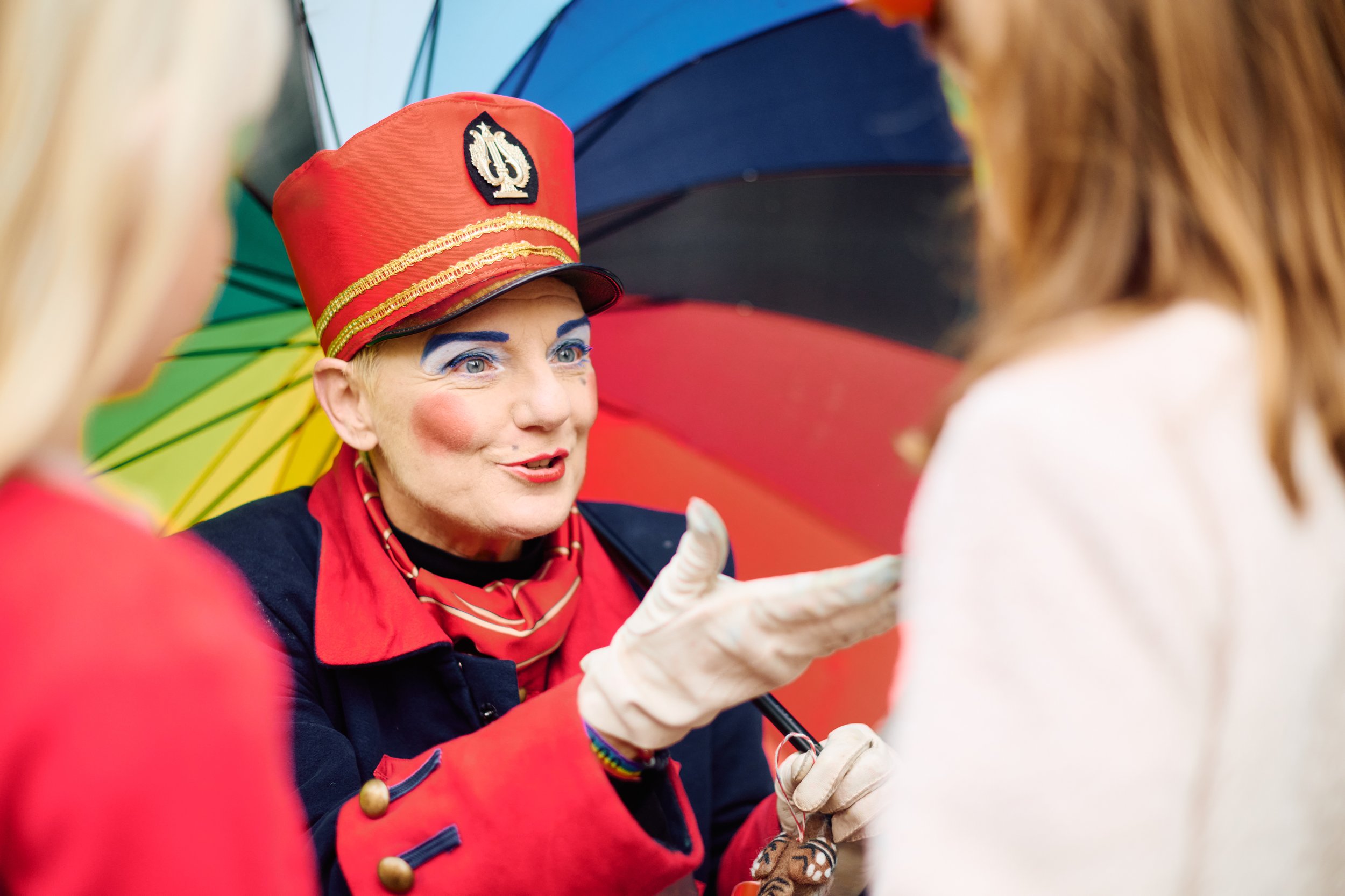 A person dressed as a circus or carnival performer, wearing a red hat with gold details, colorful makeup, and a red and navy costume, holding a small object. They are speaking to a woman with brown hair under a rainbow umbrella.