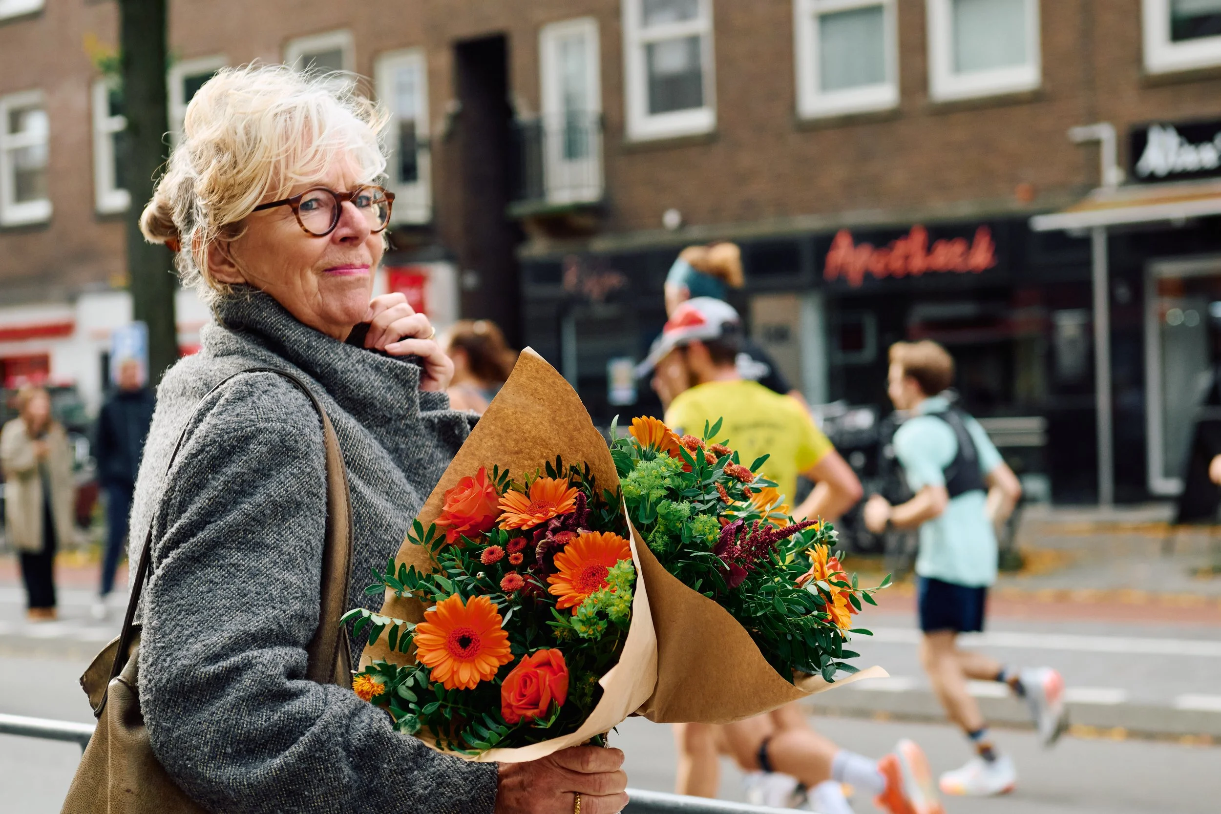 An older woman holding a bouquet of orange and yellow flowers while standing on a city street during a running event.