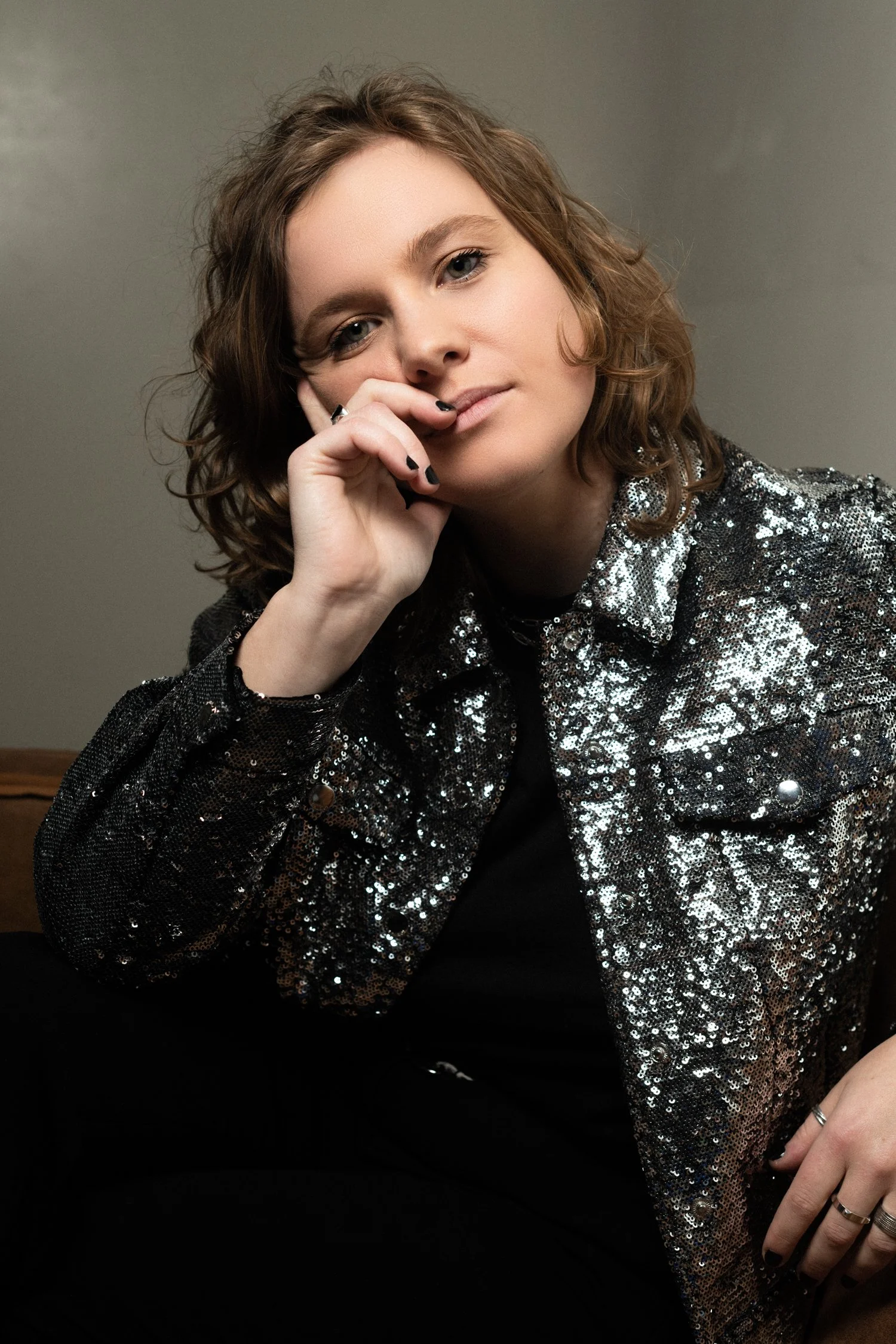 A woman with shoulder-length curly brown hair, wearing a silver sequin jacket, seated against a plain background.