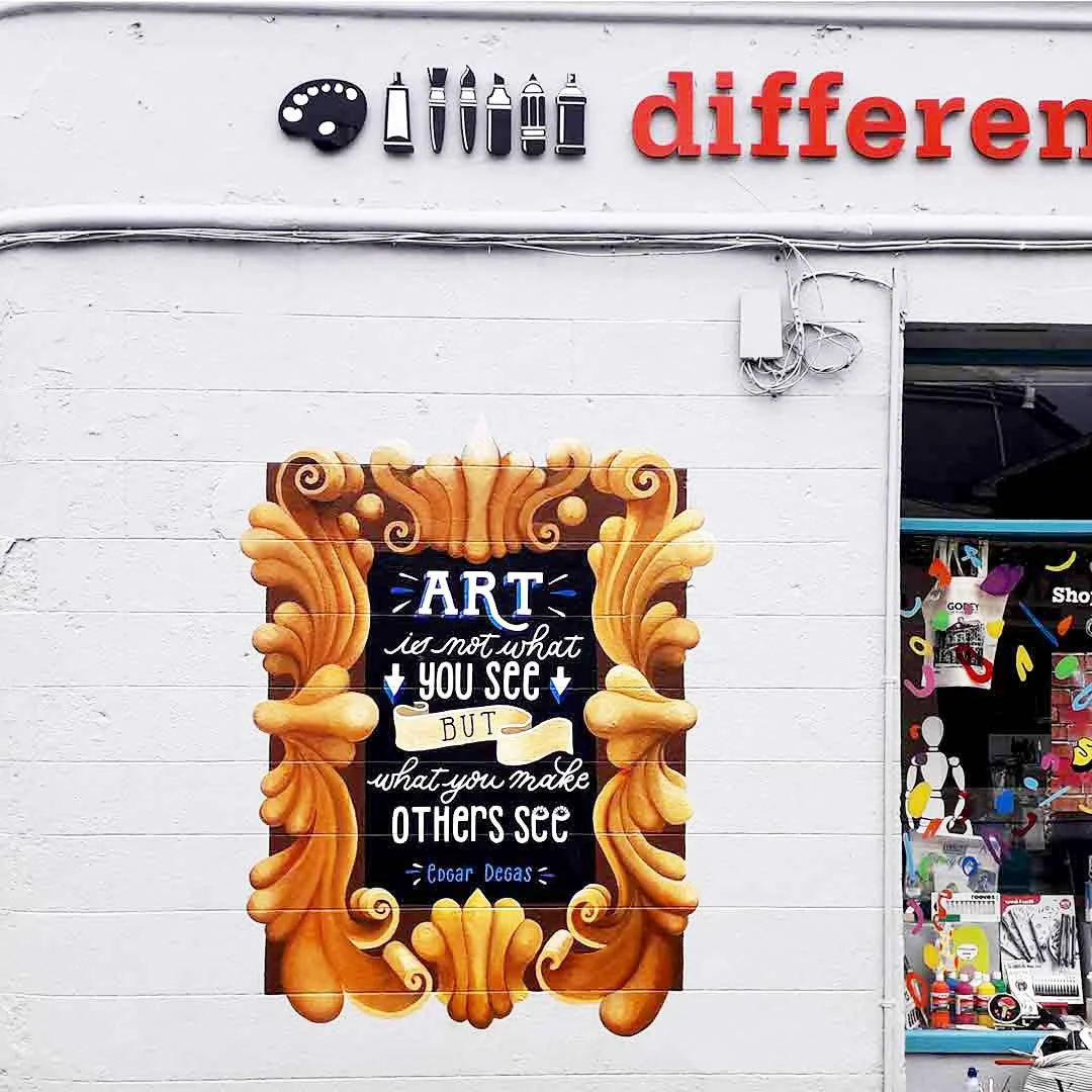Mural on an art shopfront, Different Strokes, in Gorey, Co. Wexford, Ireland. The mural features a large-scale gold vintage frame surrounding an illustrated, hand-lettered quote painted in white with blue 3D text shadows on a black background. The qu