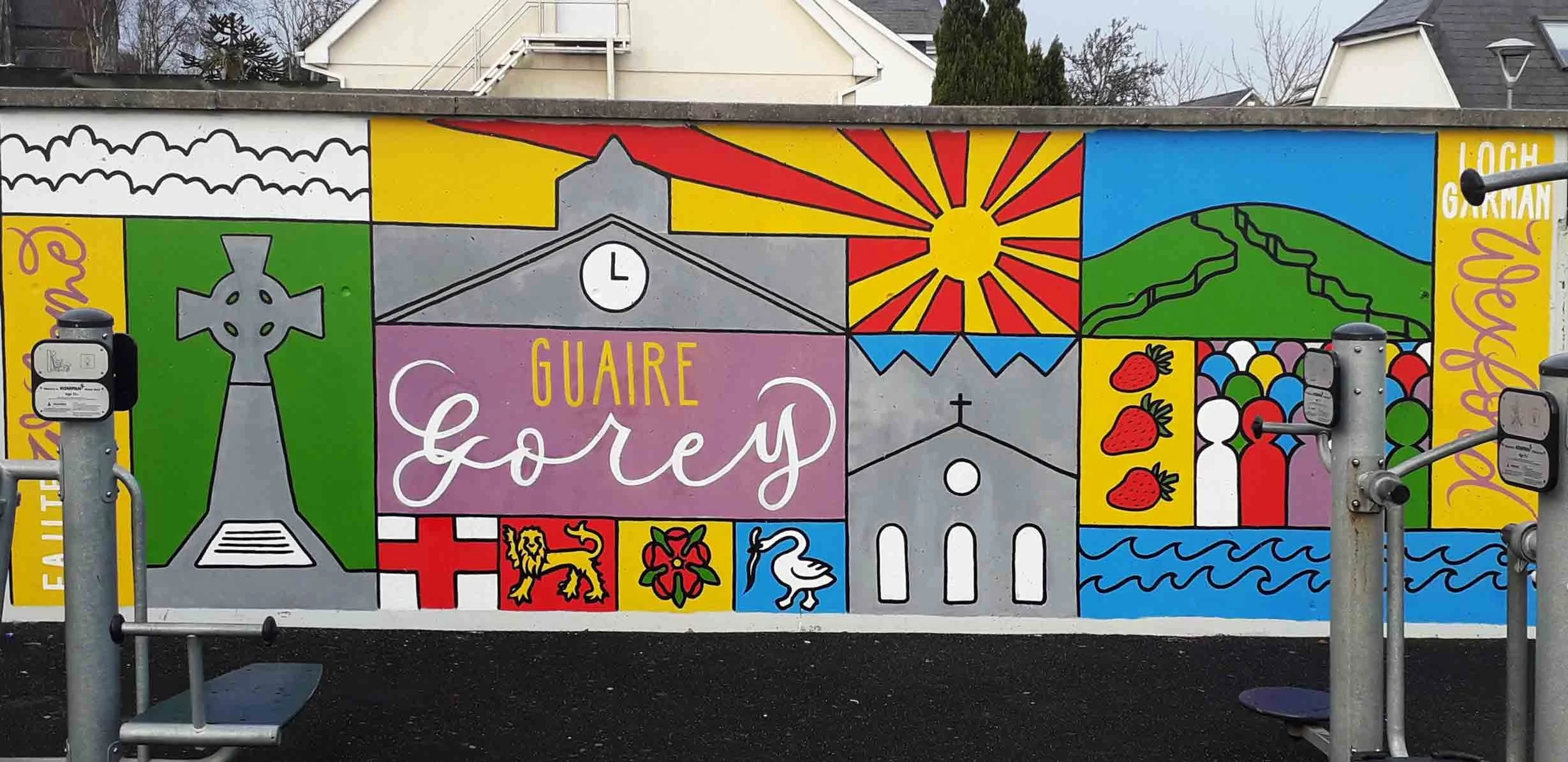 Bold, bright mural at Gorey Civic Centre featuring hand-painted local landmarks in a graphic grid-like style, hand-lettered town name and county in English and Irish in two styles of hand lettering.
