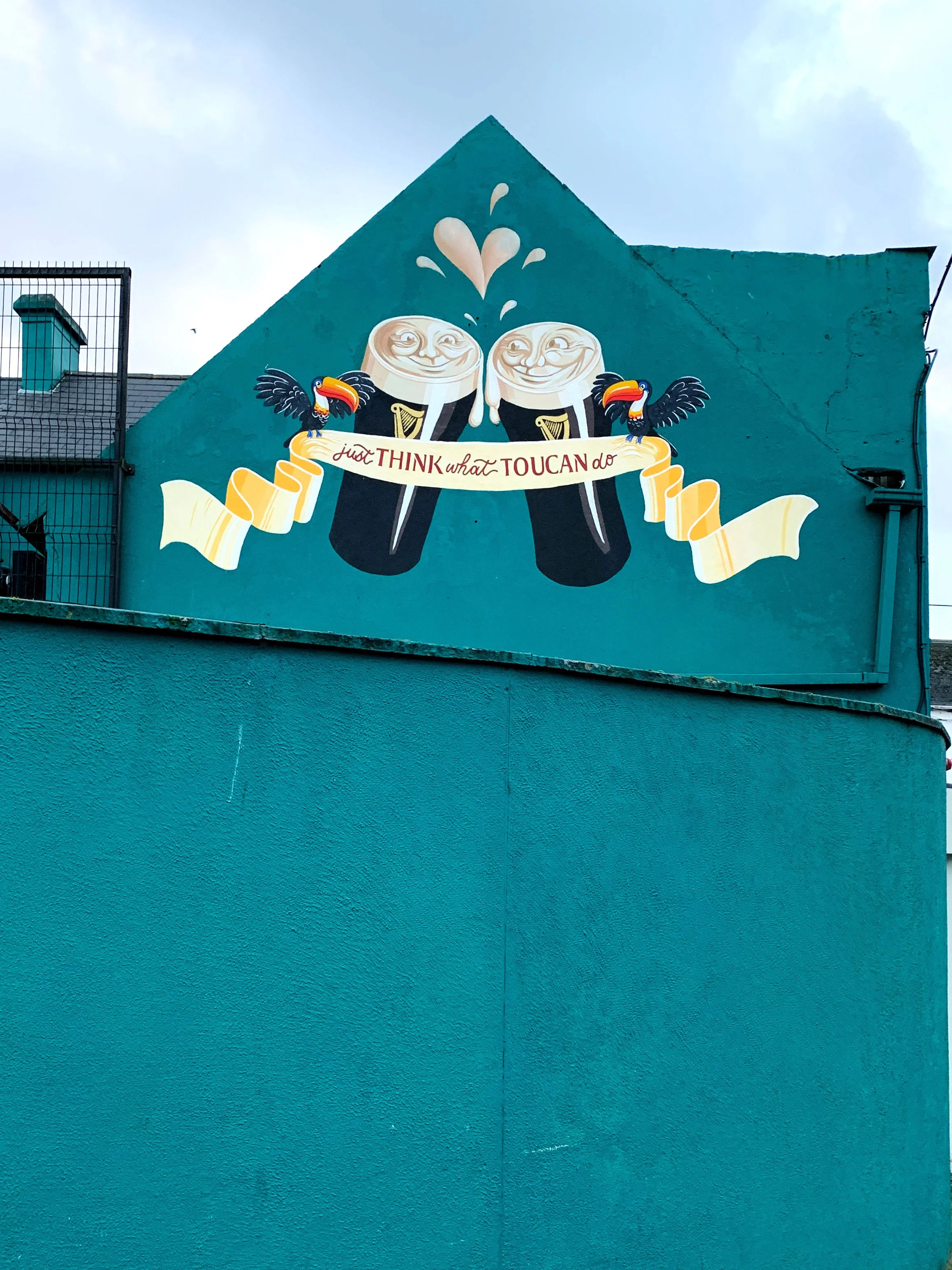 Mural on the gable end of The Brook House pub in Arklow, featuring hand-painted colourful toucans, pints of Guinness, and the slogan ‘just think what toucan do’ in vintage advertising style.