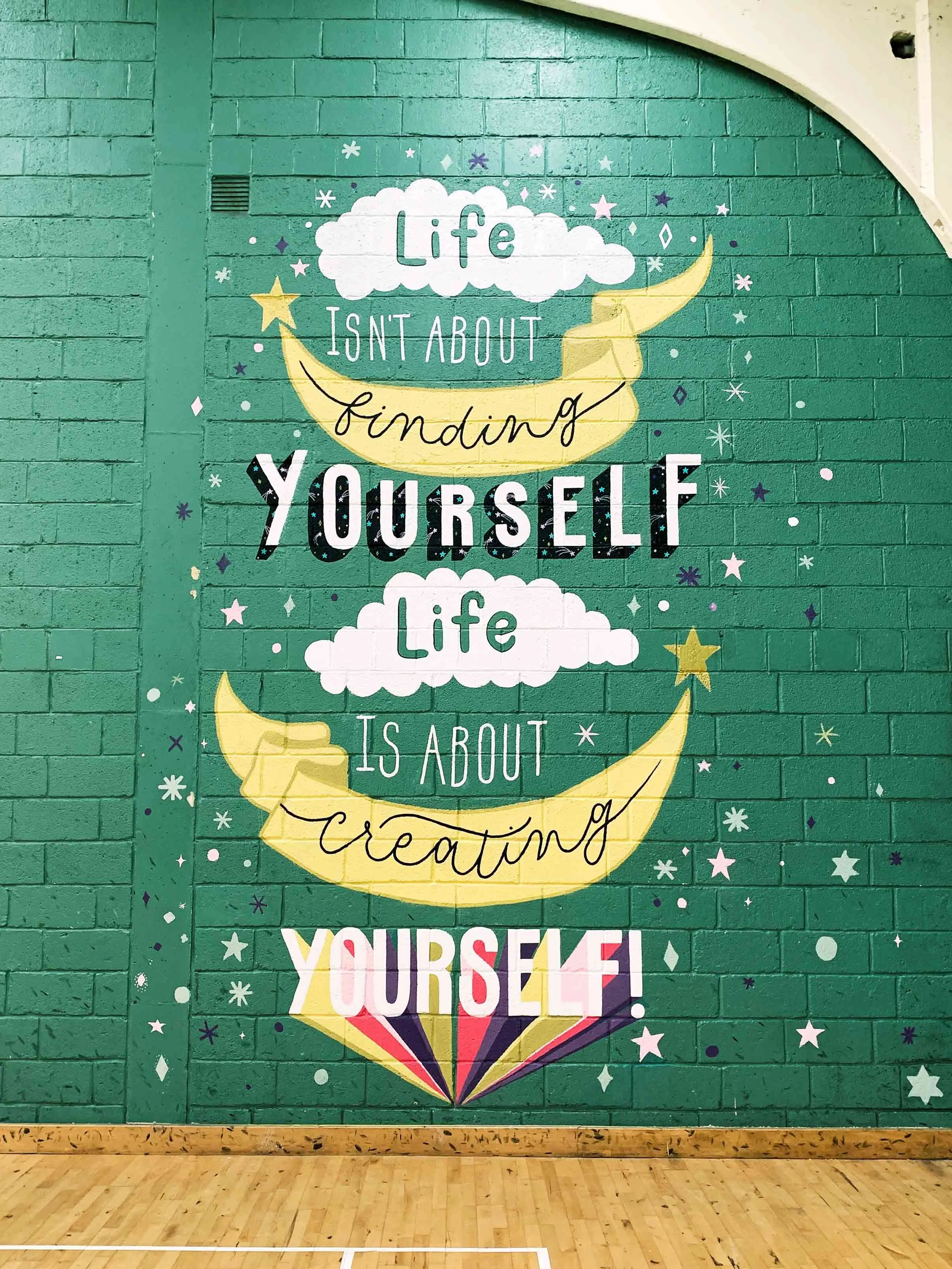 A colourful school gym mural with the quote “Life isn’t about finding yourself, life is about creating yourself” by George Bernard Shaw. The quote is written in playful, hand-lettered style, surrounded by illustrations of shooting star banners, fluff