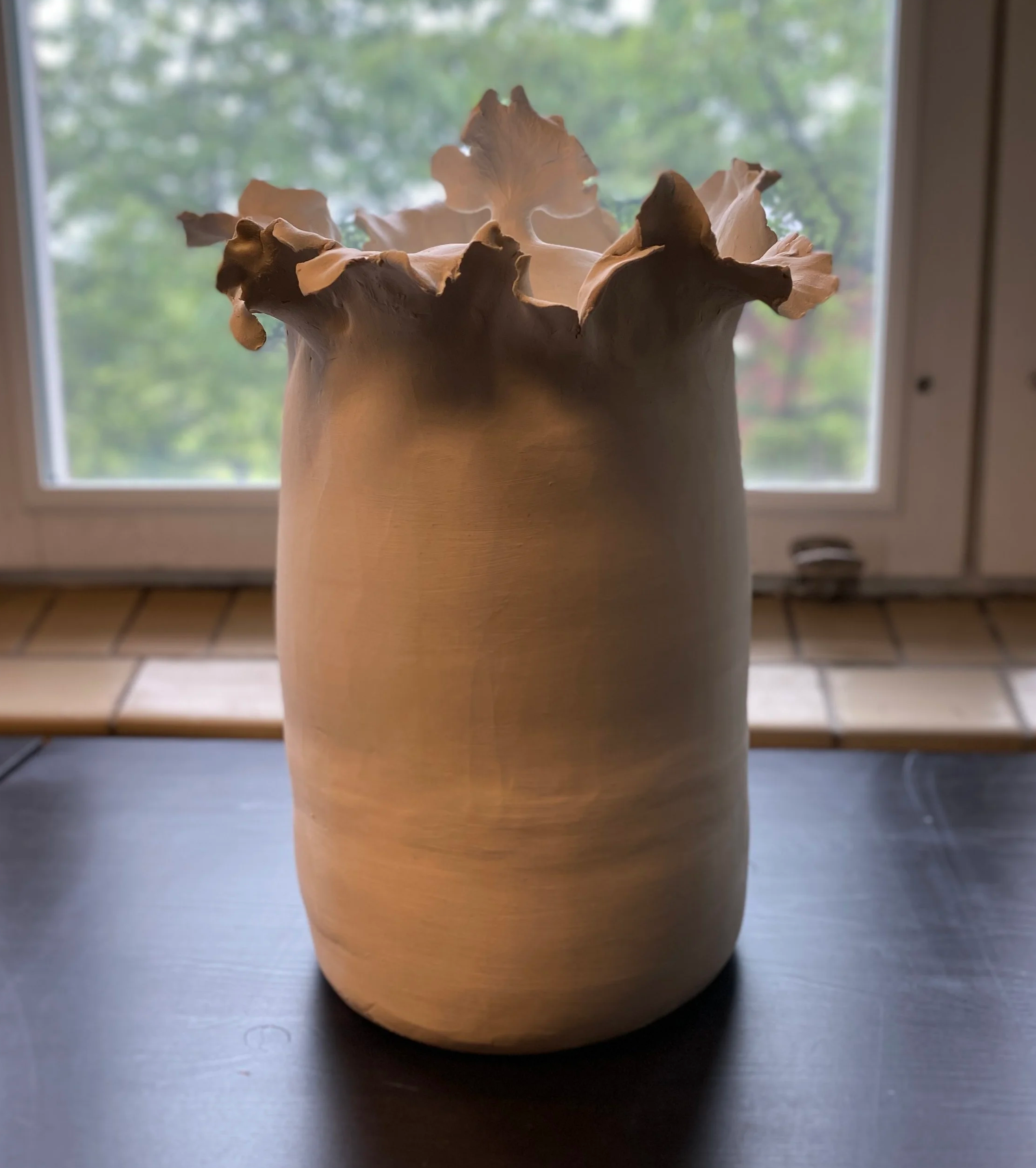 A large, smooth, beige ceramic vase with ruffled, leafy edges, placed on a dark surface near a window.