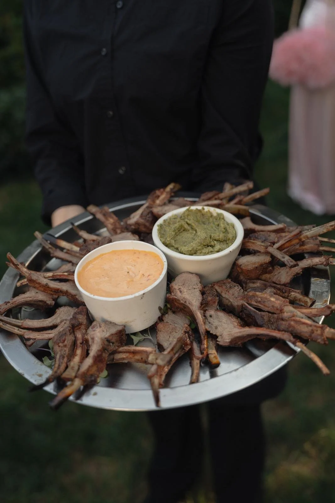 Staff of Odeen's Catering in black shirt holding a tray with grilled lamb chops, two bowls of sauce or dip, one green and one orange.