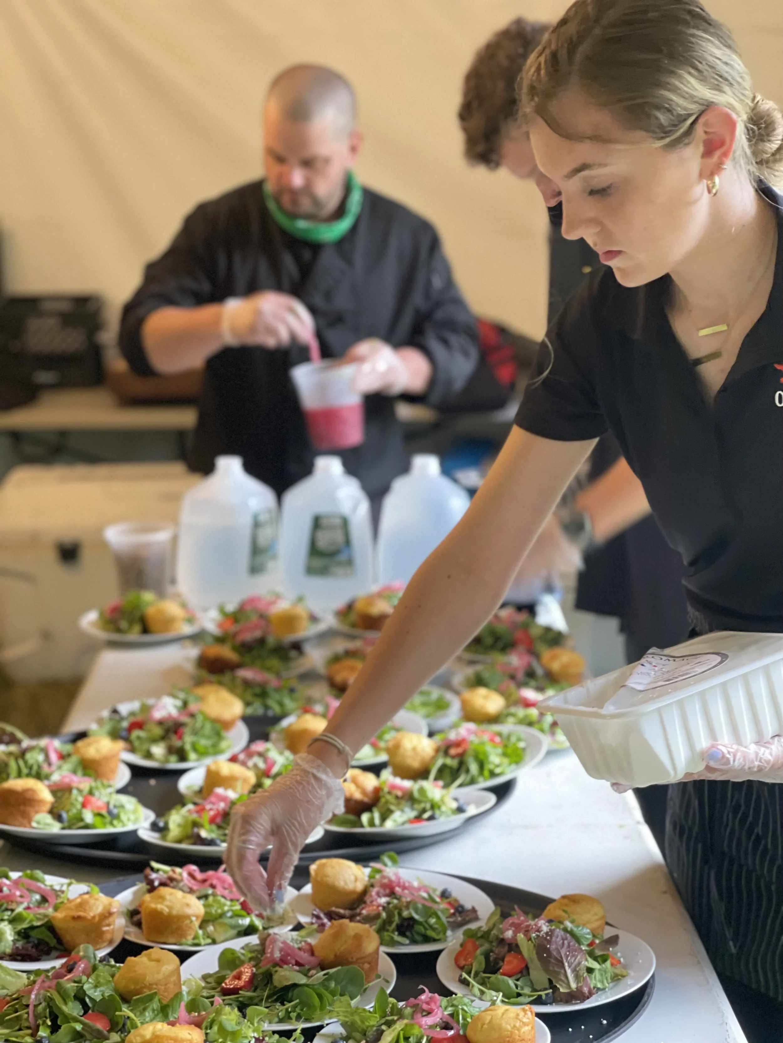 People preparing and serving salads at a catering event with plates of salad and other appetizers on a table.