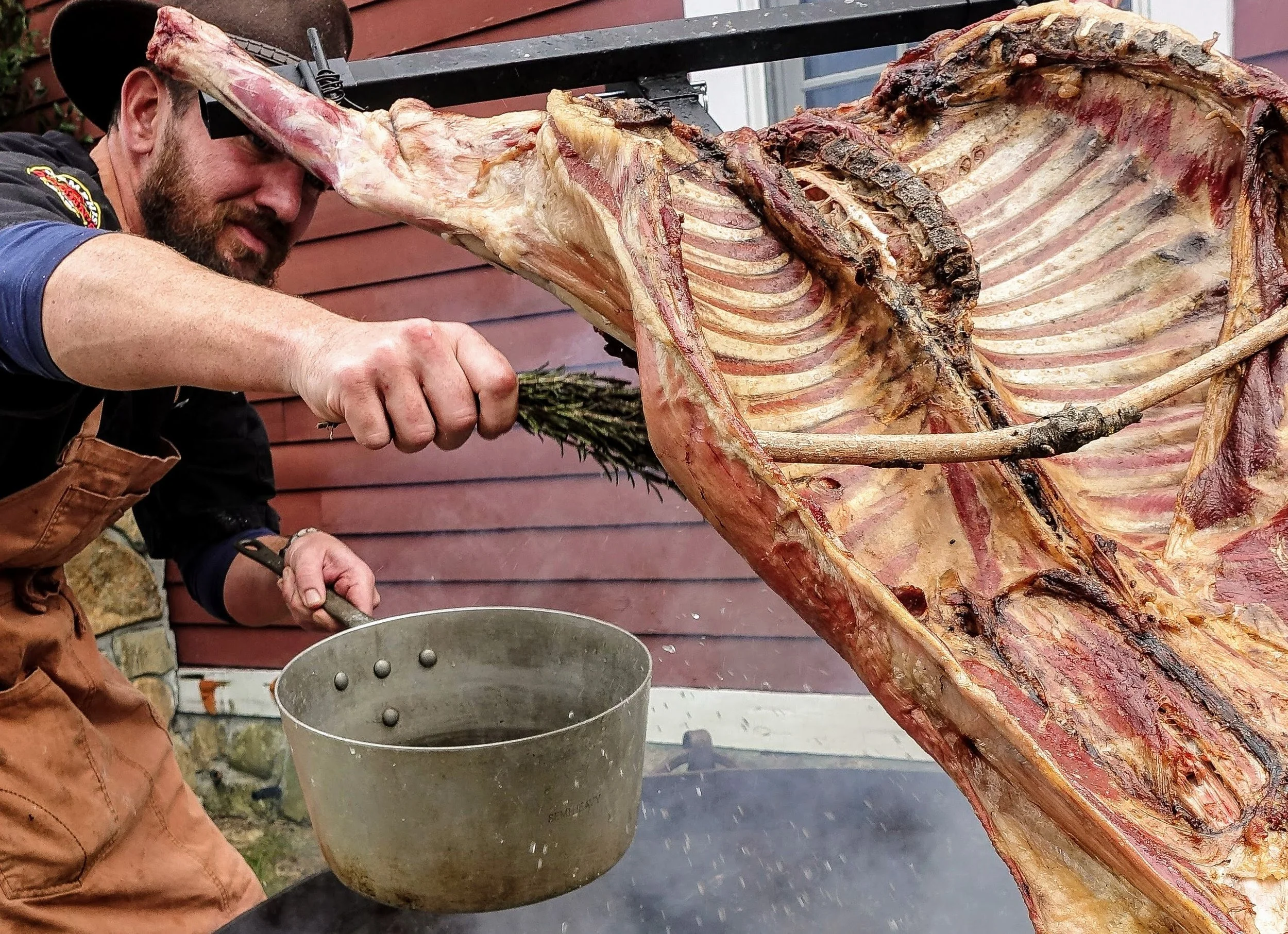 Chef basting whole lamb with rosemary sprig, open-fire BBQ