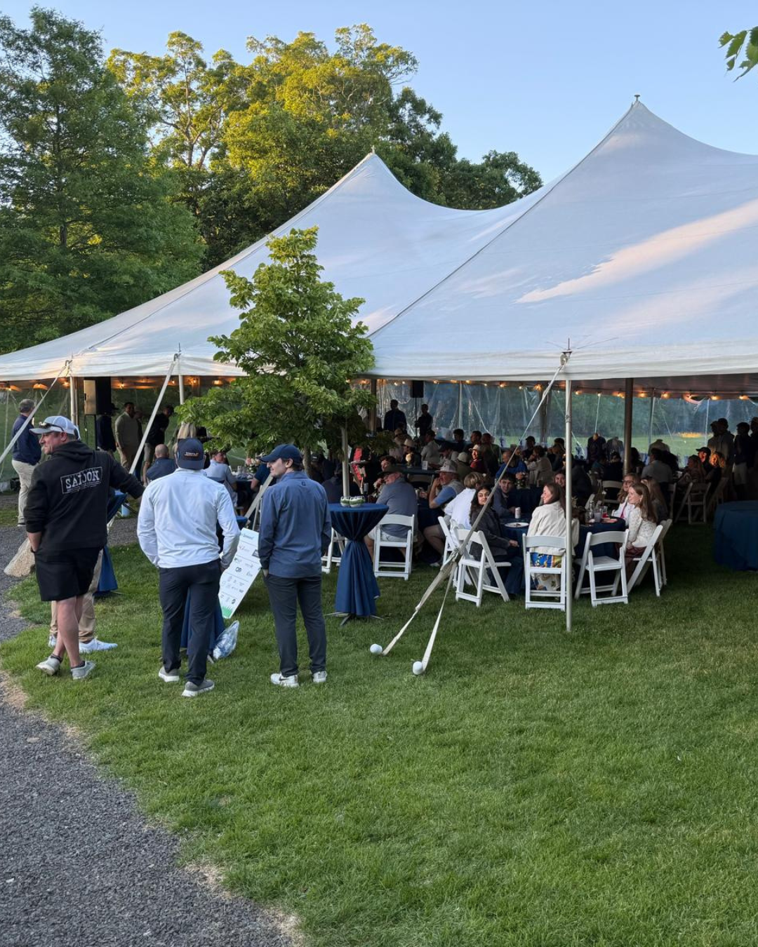 People gathered under a large white event tent outside during the evening, with some seated at tables and others standing, surrounded by green trees and grass.
