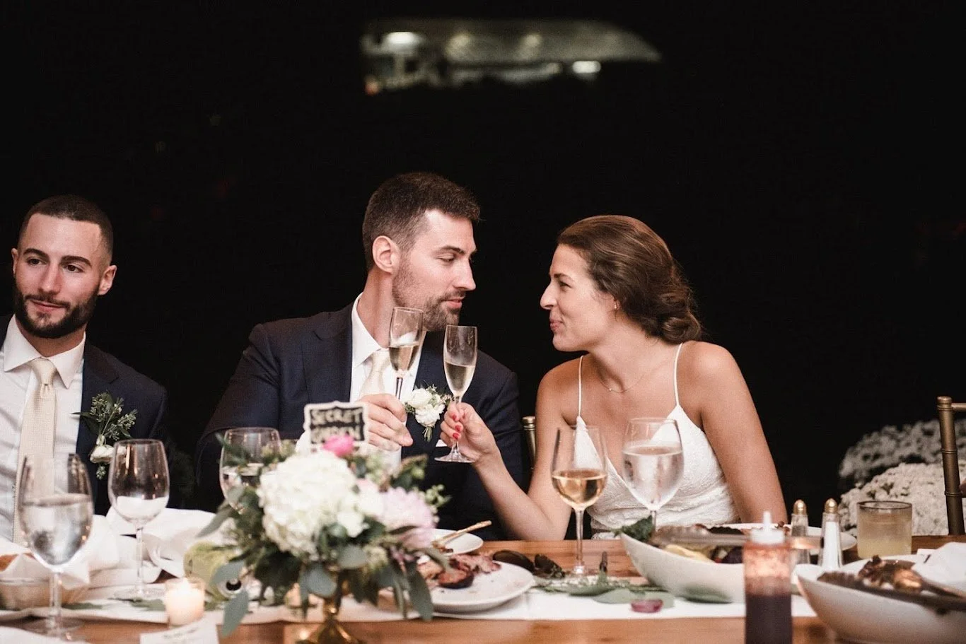 A newlywed couple at their wedding reception drinking champagne and smiling at each other, seated at a table with floral arrangements and food, with a dark background.