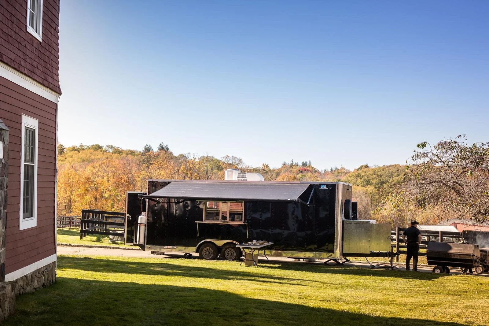 A large black food truck or trailer is parked on a grassy lawn next to a red house with white trim. There is a person standing near a barbecue grill on the right side of the image. In the background, trees with fall foliage and hills are visible under a clear blue sky.