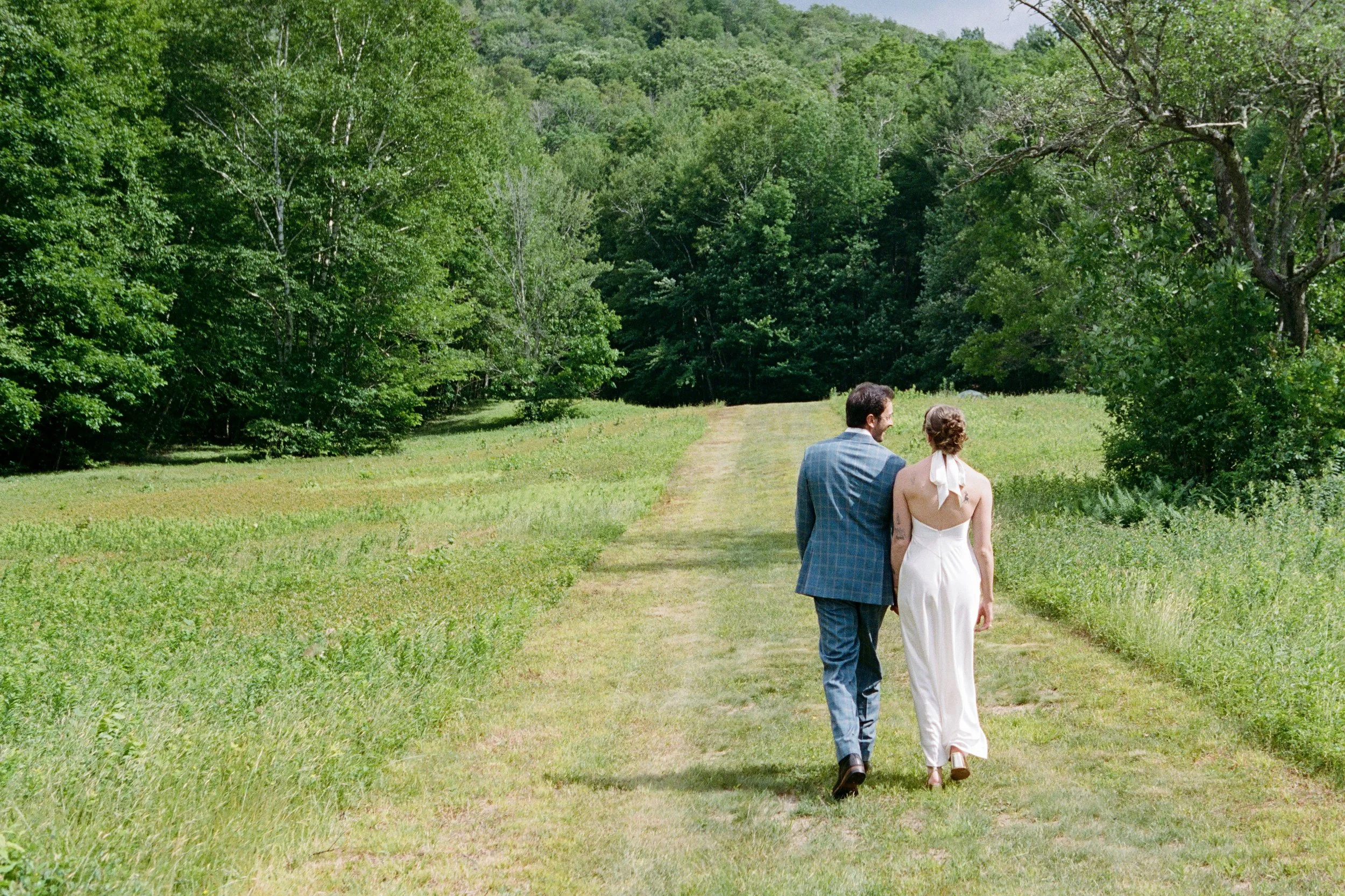 A couple walking along a grassy trail in a lush green forest, with trees on either side and dense foliage in the background.