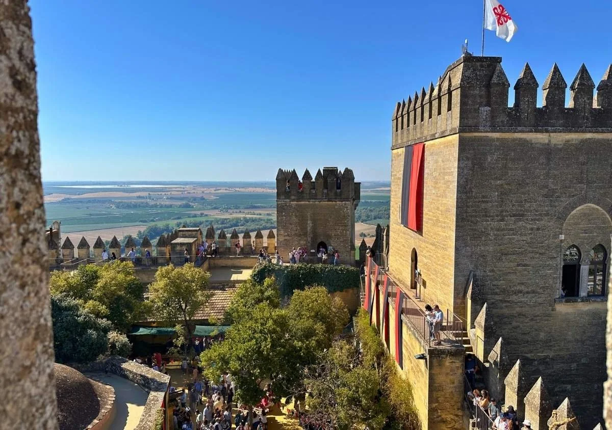 Sunlit medieval castle with stone towers and crenellated walls overlooking a wide countryside landscape under a clear blue sky, with visitors walking through the courtyard and along the ramparts.