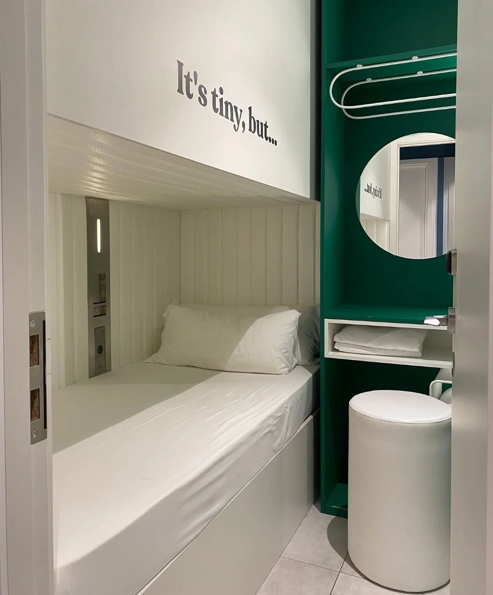 Compact single pod room at Coeo Pod Hostel in Málaga, featuring a single bed with built-in reading light and charging ports, plus an open wardrobe with shelves, mirror and stool in a modern white and green design.