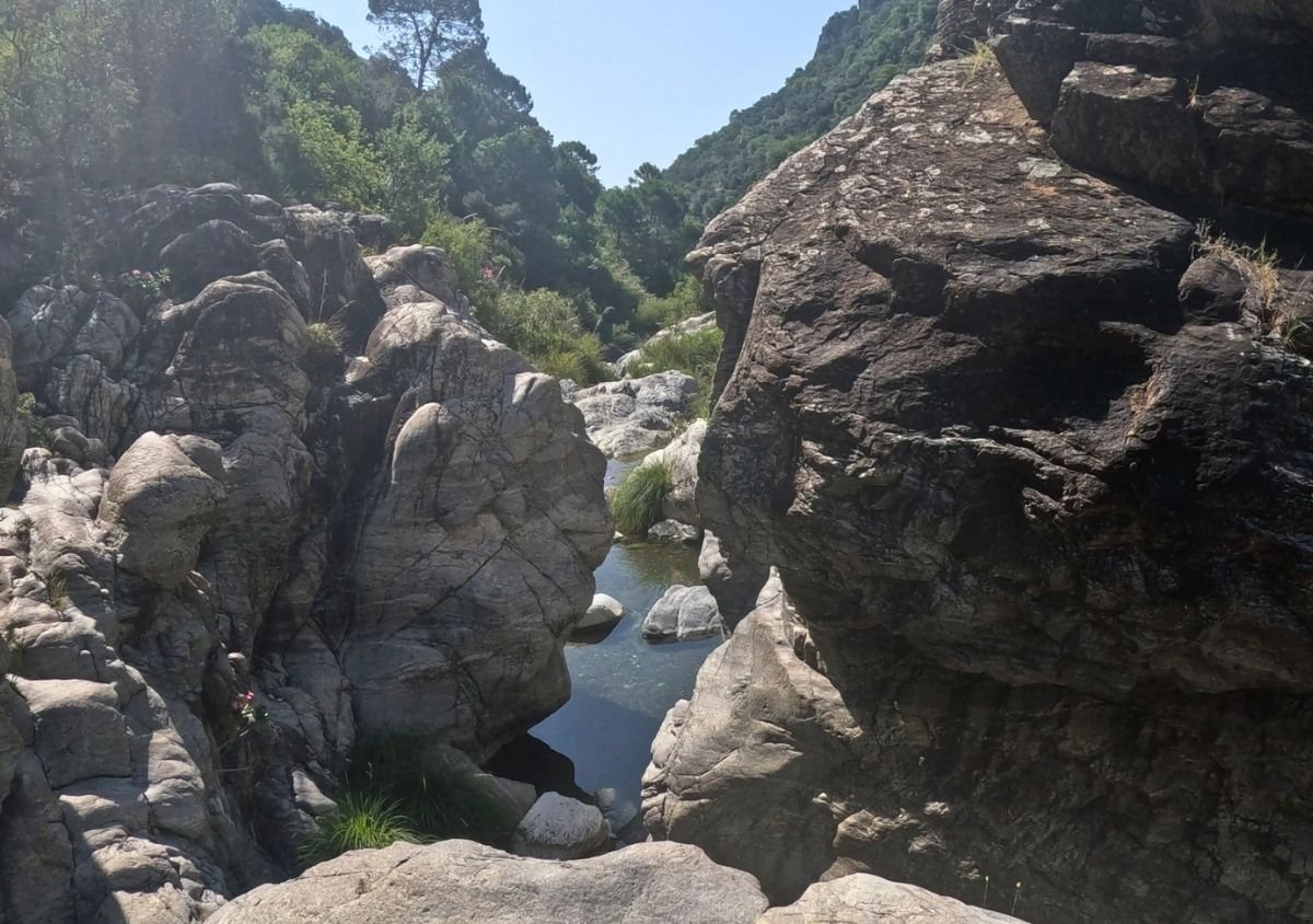 Rocky gorge along the Guadalmina River, with a small pool of clear water surrounded by large weathered boulders, grasses and tree-covered hills in the background.