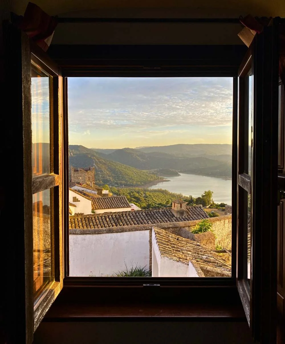 View through an open window over whitewashed village rooftops, a lakeside valley and forested hills glowing in soft golden evening light.