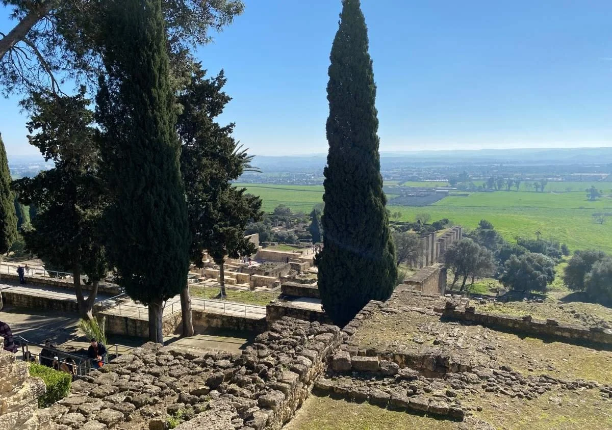 View over the ruins of Medina Azahara, with cypress trees framing the terraced archaeological site and the green Guadalquivir valley extending beyond near Córdoba, Spain.