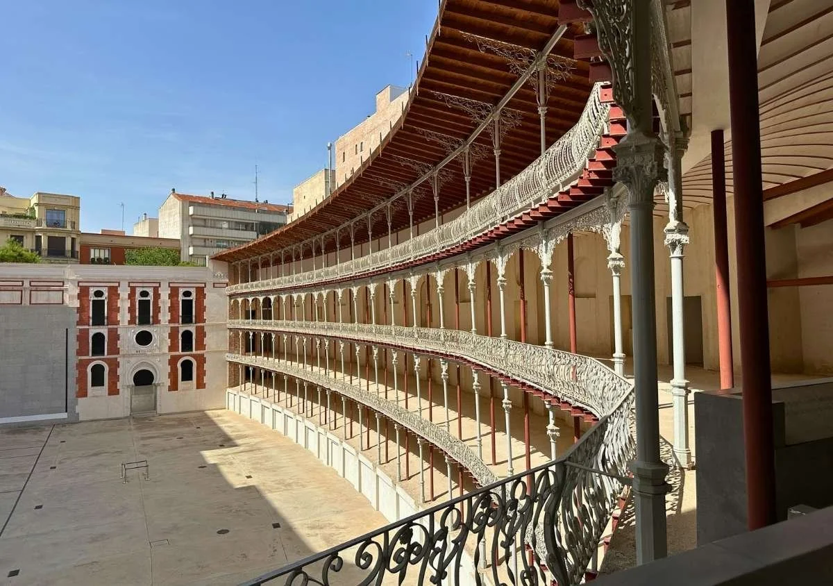 Interior view of Madrid's Frontón Beti Jai, a restored 19th-century pelota court with elegant curved, three-tiered cast-iron galleries overlooking the open playing court, illuminated by bright sunlight beneath a clear blue sky.