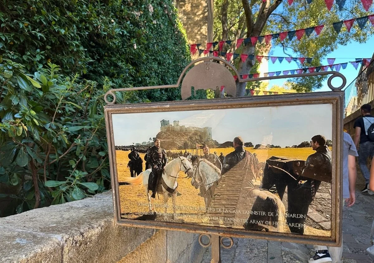 nformation panel at Almodóvar del Río Castle showing a Game of Thrones filming scene, with the real castle visible in the background and visitors walking along the shaded pathway beneath colourful bunting.