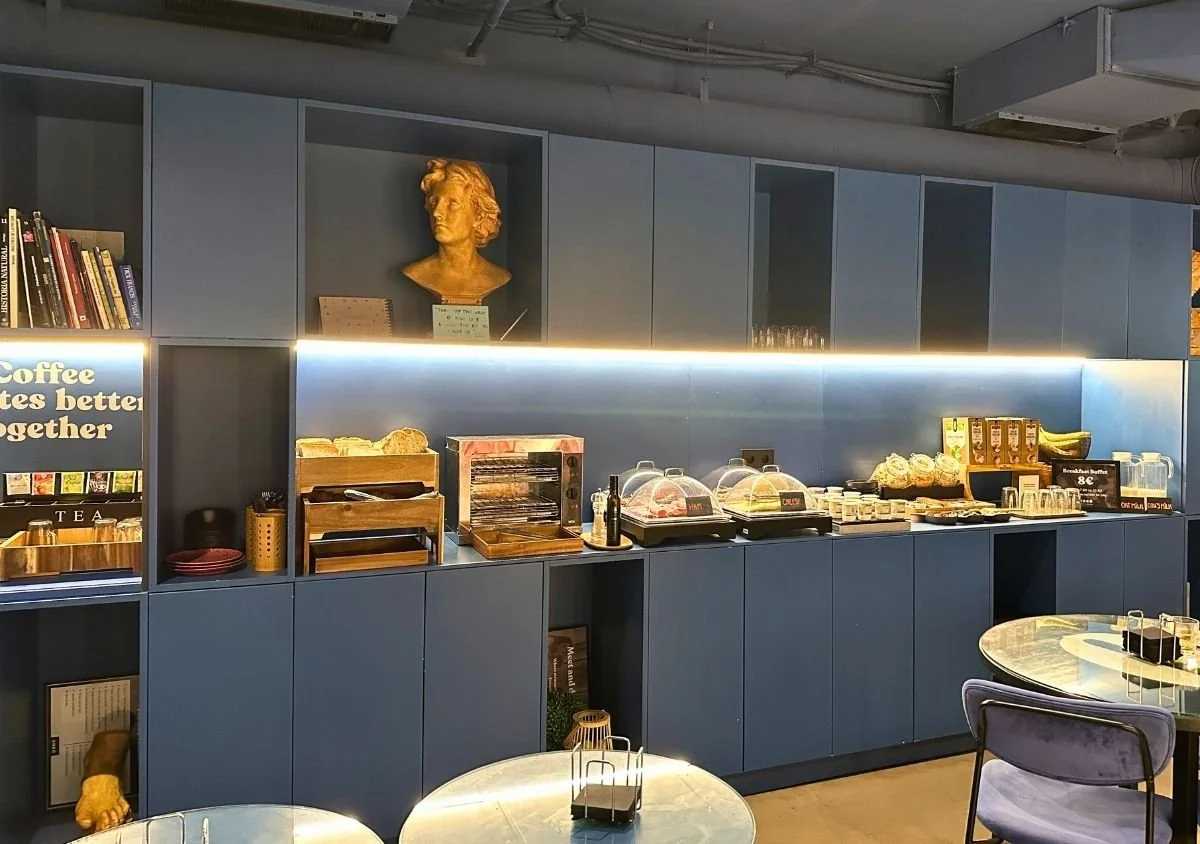 Breakfast buffet at Coeo Pod Hostel in Málaga, with fresh bread, pastries, cereal, yoghurt, fruit, juice and hot drinks displayed on a modern blue counter beneath softly lit shelving and decorative sculptures.