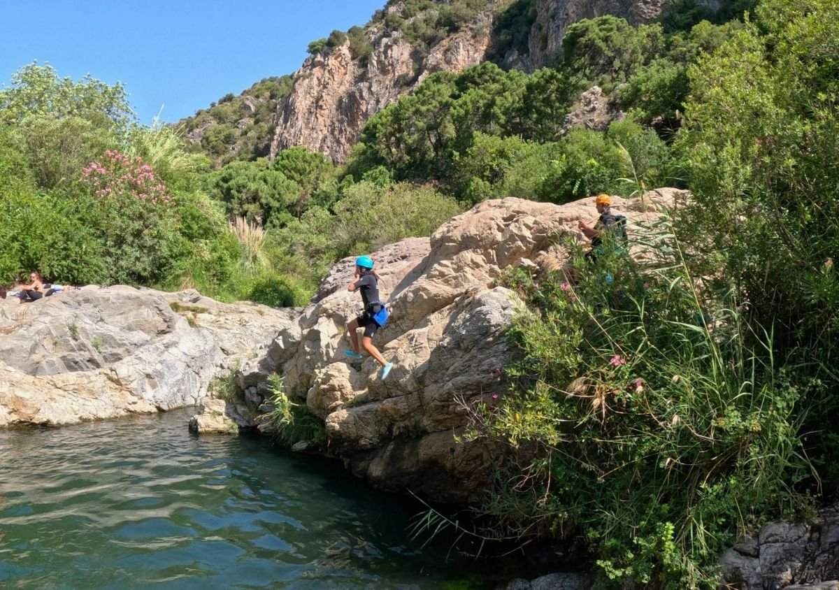 A canyoner in a blue helmet jumps from a rock beside a natural pool in the Guadalmina River canyon, surrounded by lush vegetation and rocky cliffs.