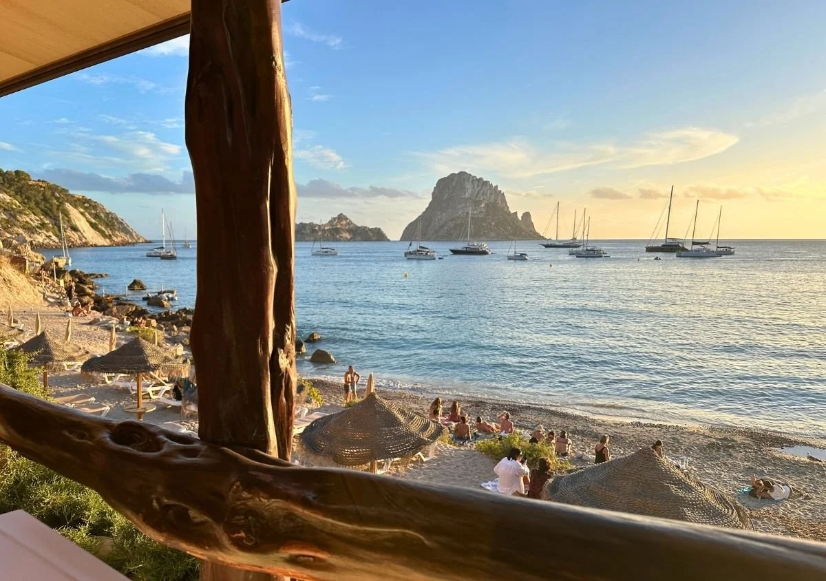 View of Es Vedrà from Restaurante El Carmen, with sailboats on the sea and a beach lined with thatched parasols