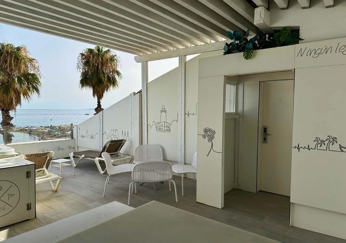 Shaded terrace at La Chancla Hotel in Pedregalejo, Málaga, with white lounge chairs overlooking the Mediterranean Sea and beach, framed by palm trees and minimalist white décor.