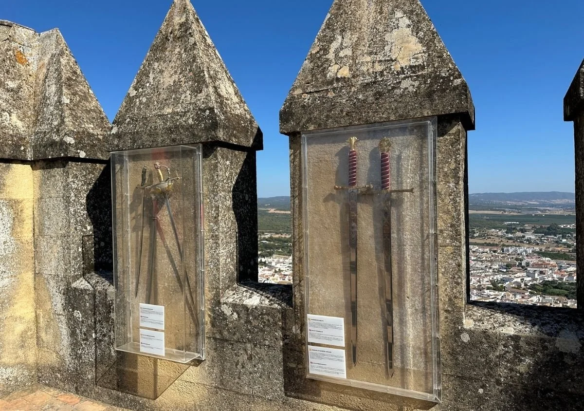 Historic swords displayed in protective cases along the battlements of Almodóvar del Río Castle, with panoramic views over the town and surrounding countryside beneath a clear blue sky.